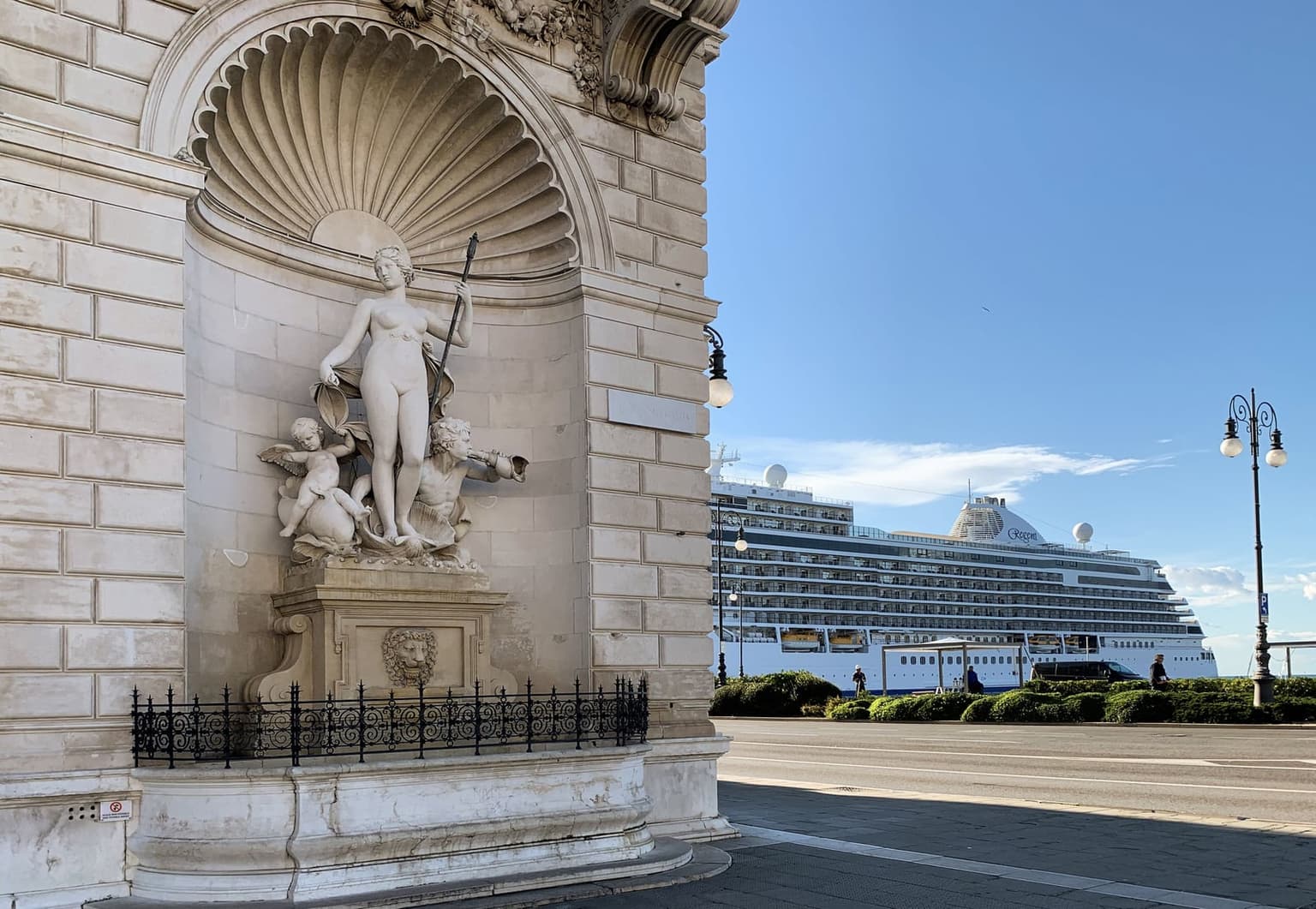A marble fountain of a goddess with cherubs in a shell-shaped niche on a stone seafront palace in Trieste, with a large white cruise ship berthed just across the waterfront road