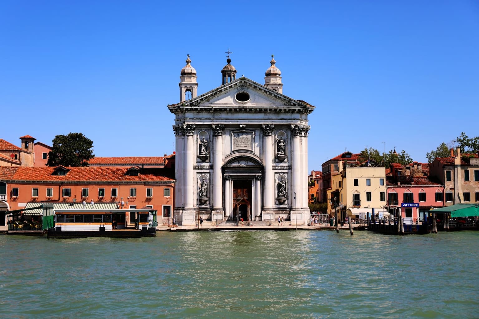 The white columned facade of the Gesuati church on the Zattere waterfront, seen across the Giudecca Canal between orange and pastel houses, with a vaporetto pontoon at left and a blue Zattere stop sign at right