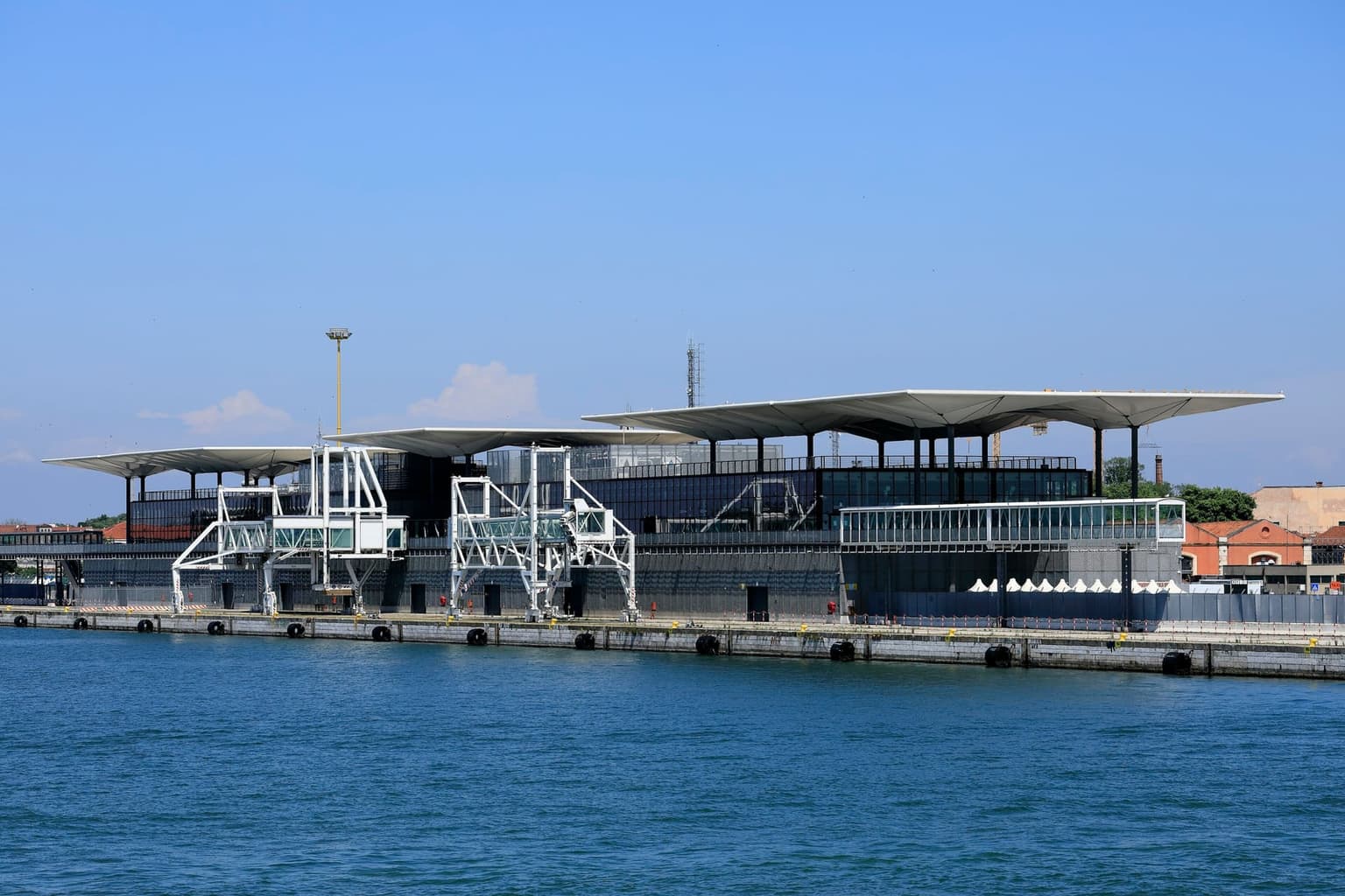 The Marittima cruise terminal in Venice seen from the water, a low dark-glass building under white umbrella-shaped canopies, with two white boarding bridges on an empty quay and no ship alongside