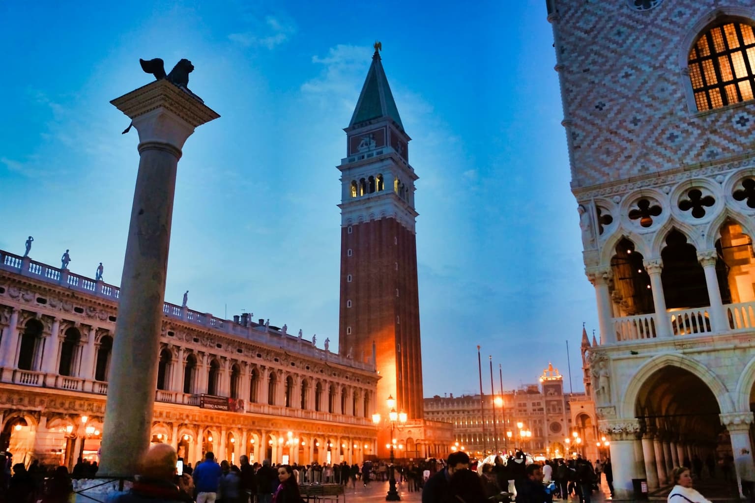 The Campanile of San Marco floodlit against a deep blue evening sky, seen from the Piazzetta with the winged-lion column at left, the lit arcades of the Marciana library below, and the corner of the Doge's Palace at right