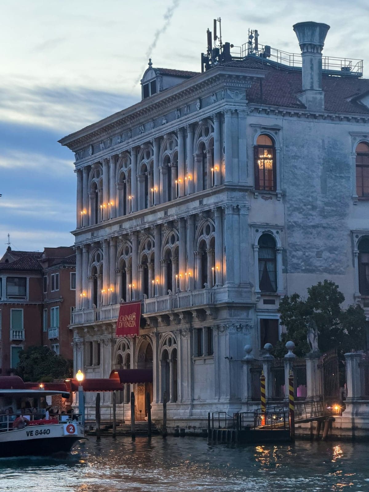 The marble facade of Ca' Vendramin Calergi on the Grand Canal at dusk, candle lights glowing in its arched windows and a red Casino di Venezia banner over the water entrance, with a vaporetto passing at left
