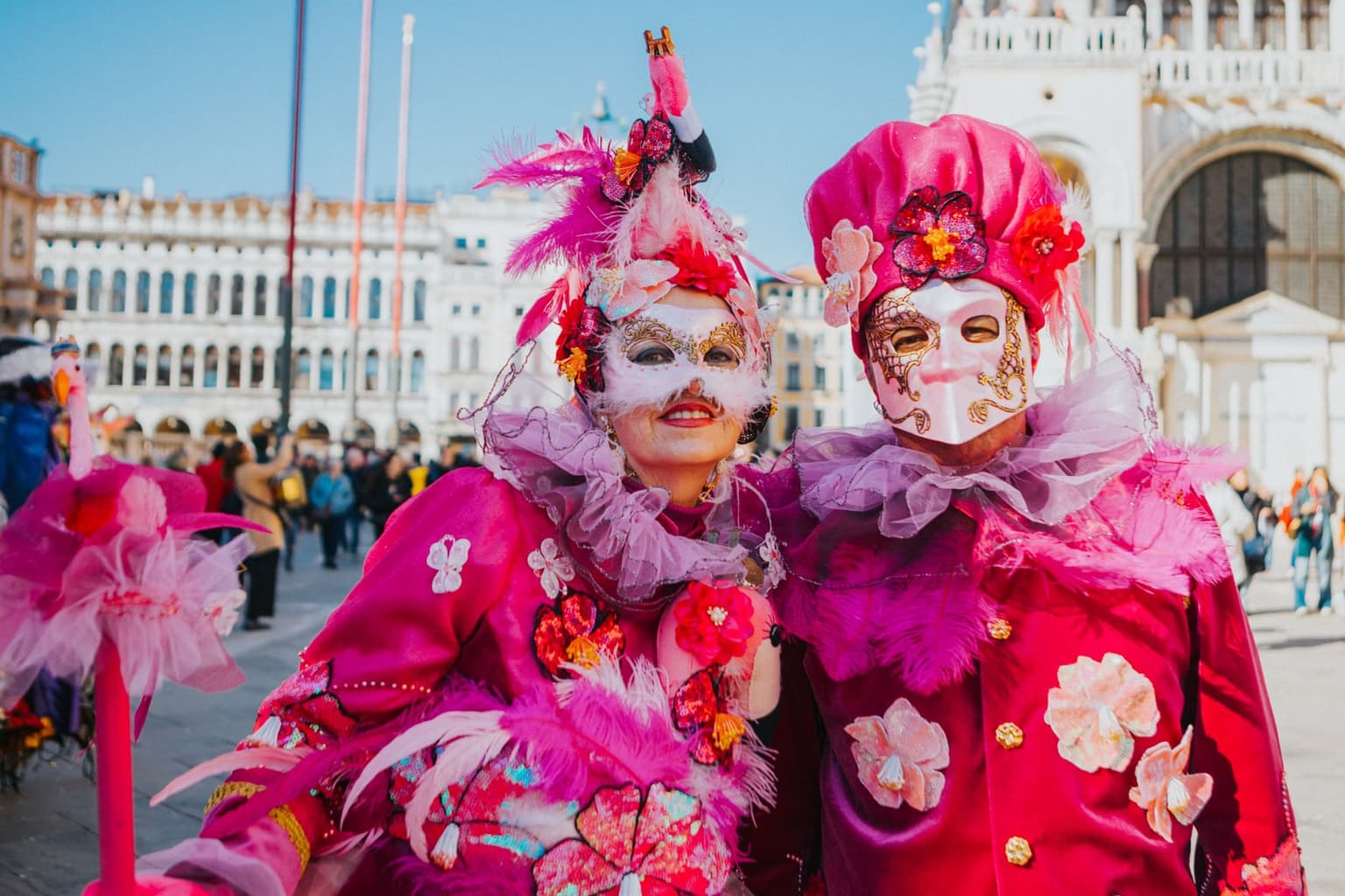Two Carnival maskers in matching pink and magenta costumes trimmed with feathers, ruffs and sequined flowers, posing in a sunny Piazza San Marco with the crowd and the arcades of the Procuratie behind them