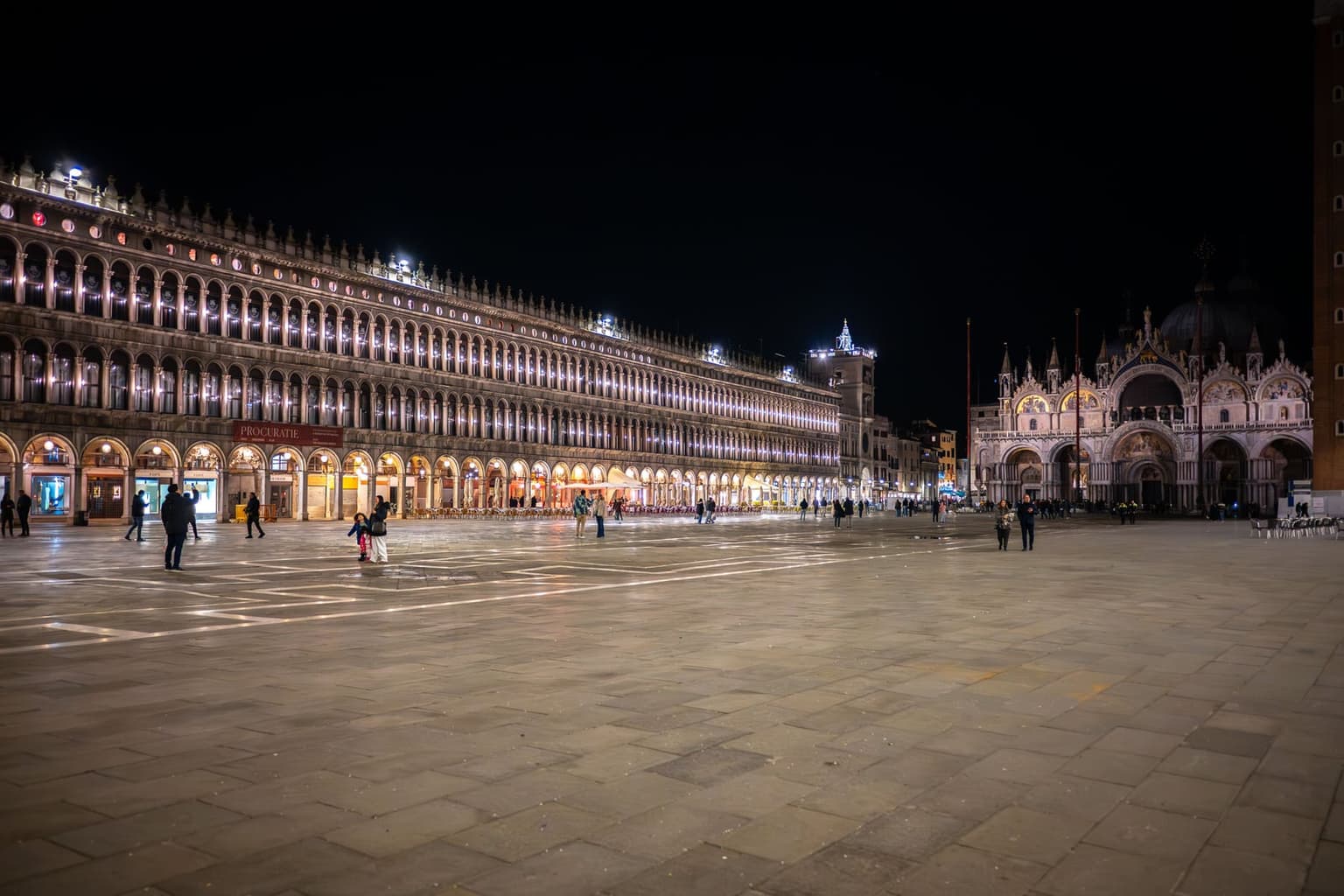 Piazza San Marco at night, almost empty, with the long arcades of the Procuratie Vecchie lit along the left and St. Mark's Basilica and the Clock Tower glowing at the far end