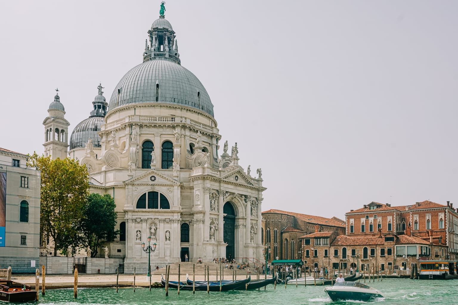 The domed basilica of Santa Maria della Salute across the Grand Canal under a hazy sky, gondolas moored by its steps and a motorboat crossing in front