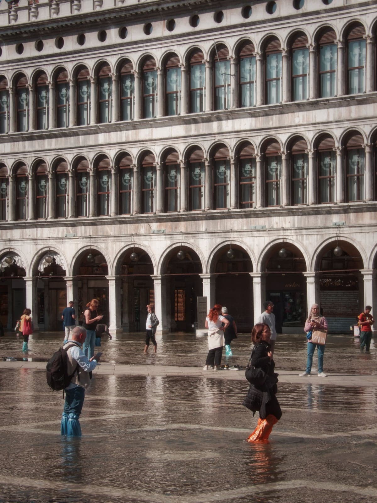Piazza San Marco under high water, visitors in plastic boot covers wading across the flooded paving in front of the arcades of the Procuratie