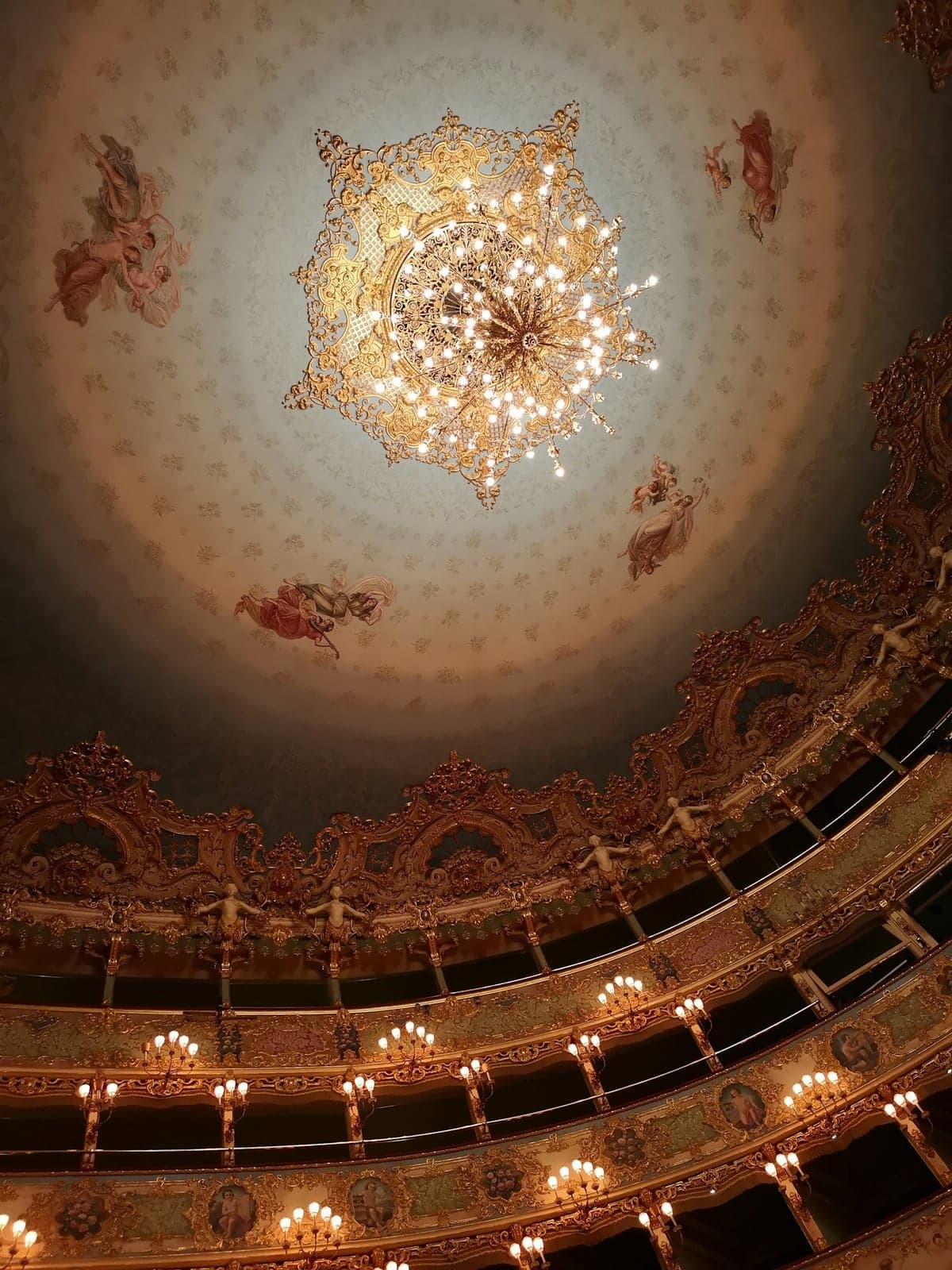 Looking up inside Teatro La Fenice at the painted ceiling with its large gilded chandelier and floating figures, above tiers of gilded opera boxes lit by candle-style lamps