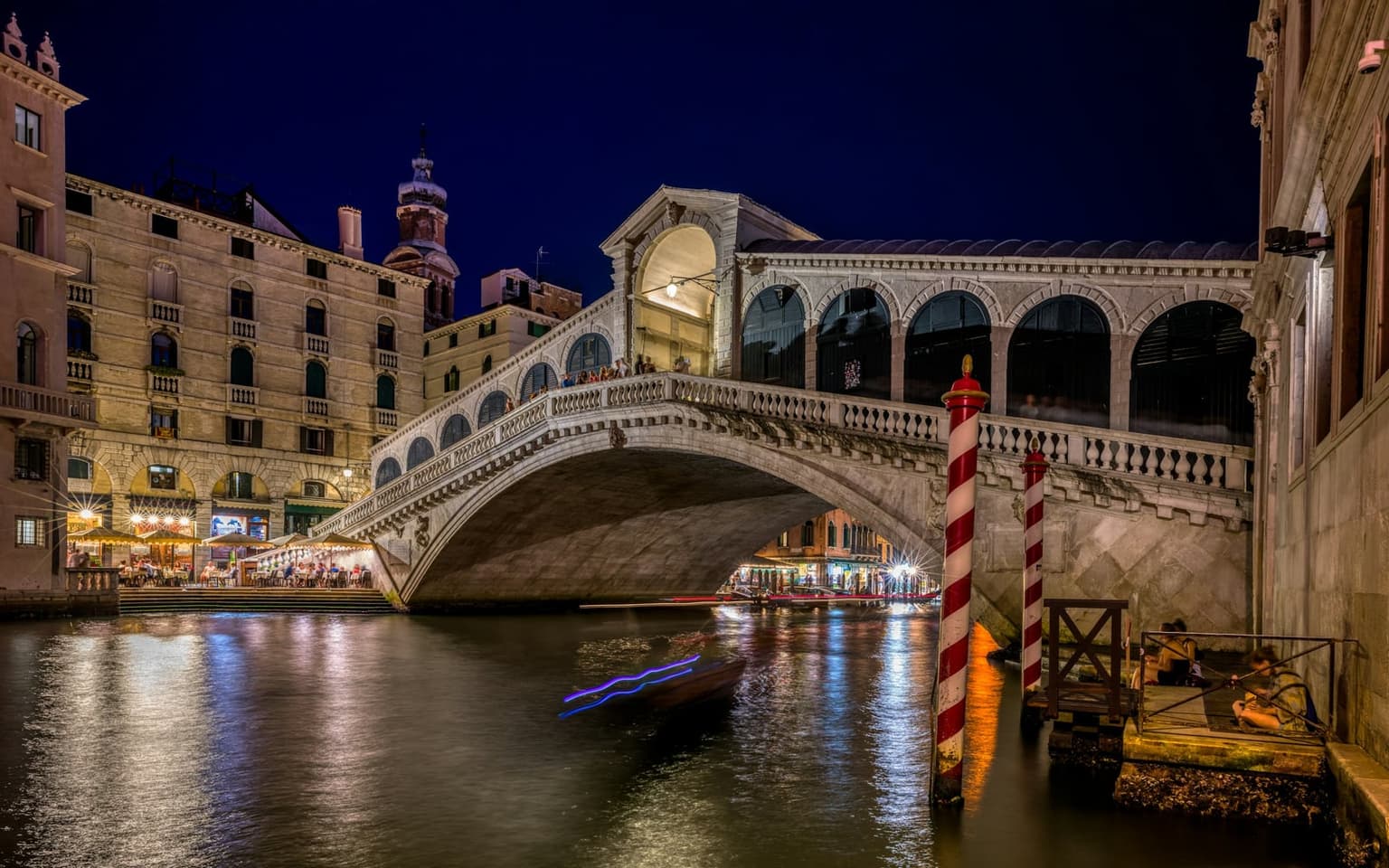 The Rialto Bridge lit at night over the Grand Canal, restaurant tables glowing along the bank at left and red-and-white striped mooring poles in the water at right