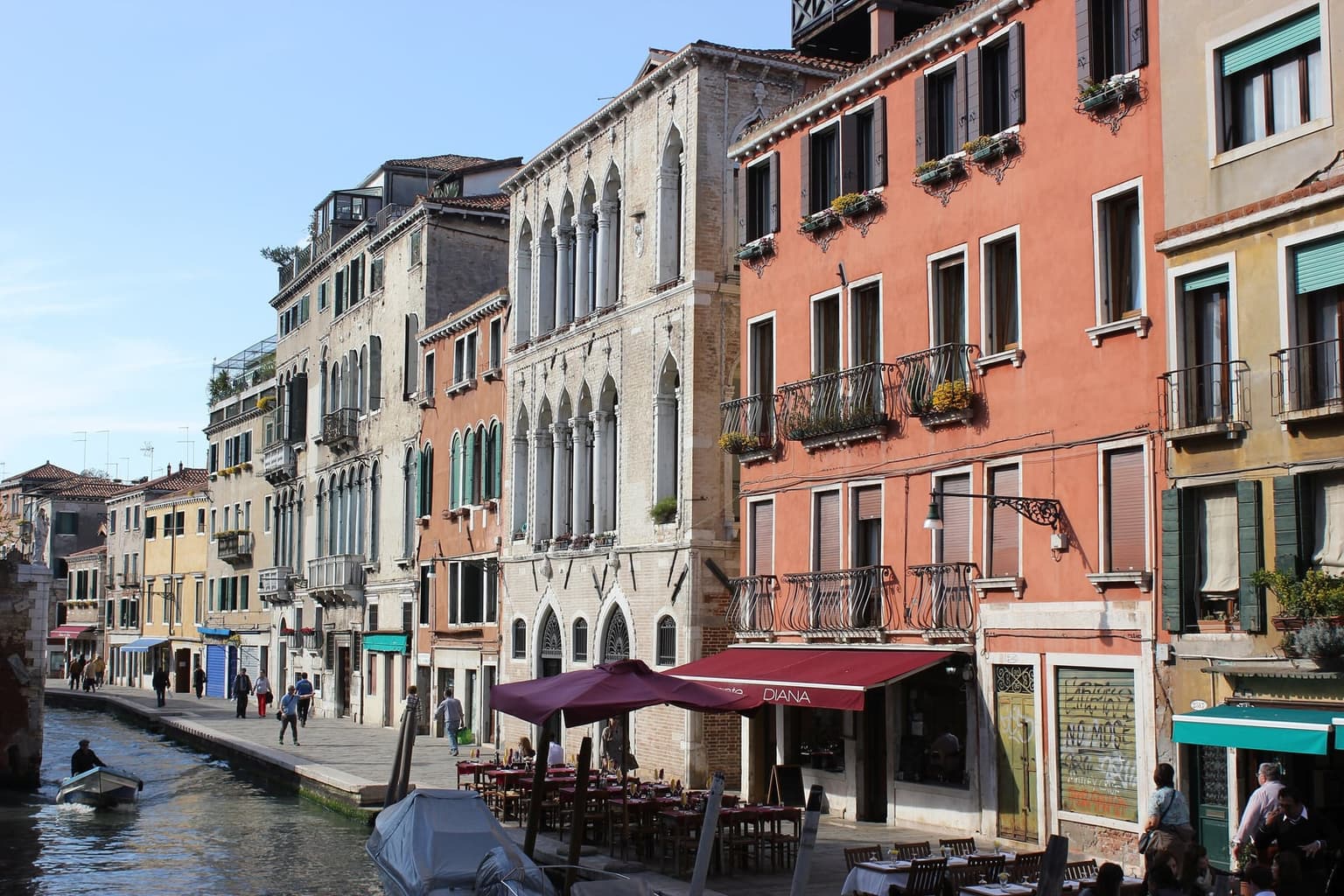 A canal-side walkway lined with pastel houses and a Gothic palazzo, restaurant tables and burgundy umbrellas set out along the water, and a small motorboat on the canal