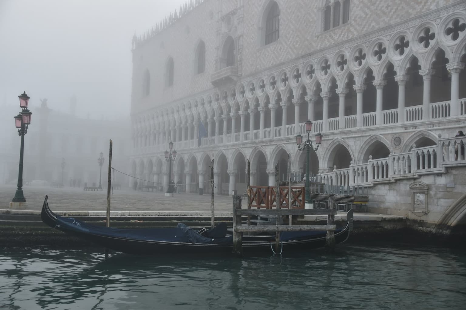 A covered gondola moored in front of the Doge's Palace in thick fog, the Piazzetta behind it almost empty, with pink-globed lampposts and the winged-lion column fading into the mist