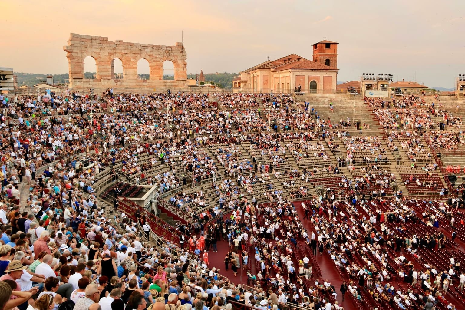 A large audience fills the stone steps and the rows of red seats inside the Arena di Verona at sunset before an evening performance, with the four arches of the surviving outer wall rising against a pink sky