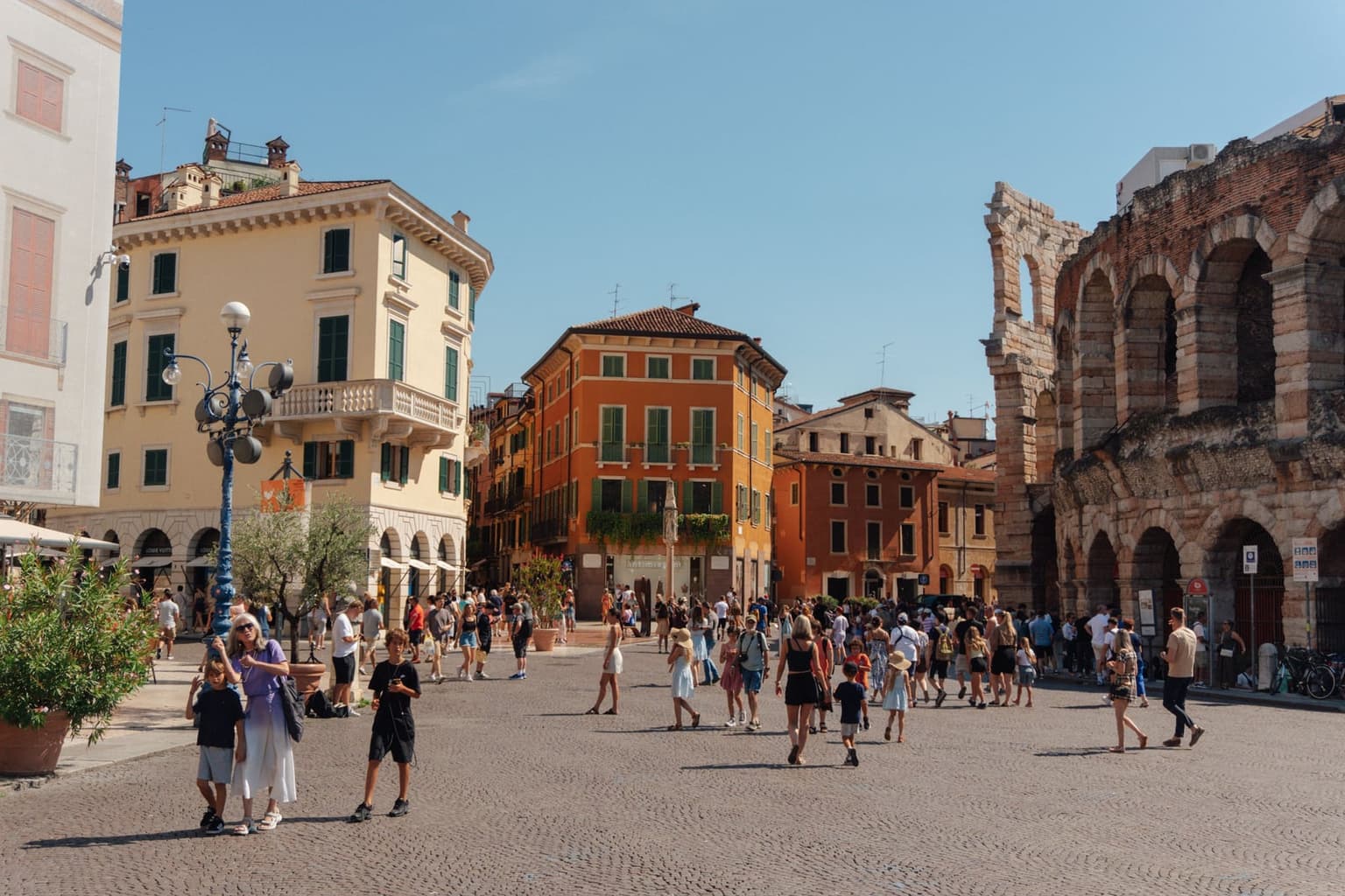 Visitors cross the cobbled Piazza Bra in Verona on a sunny summer day, with the two-story stone arches of the Arena on the right and cream and orange palazzi with green shutters across the square