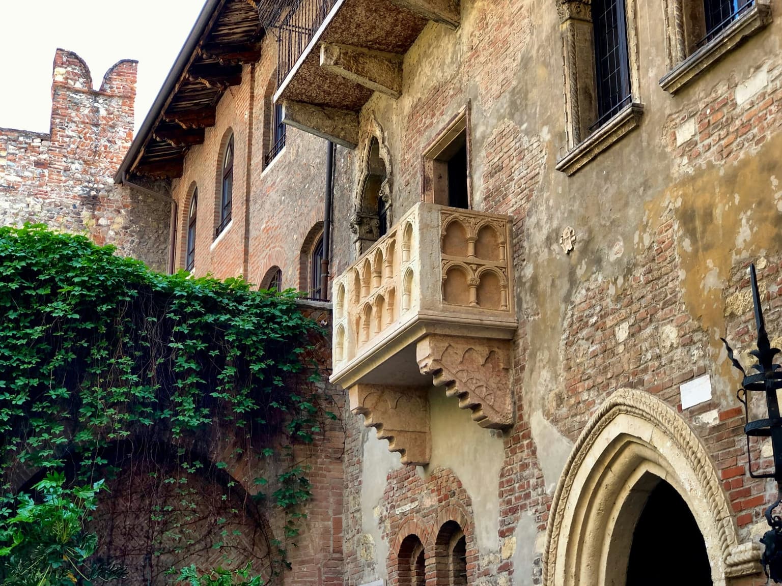 The carved stone balcony of Juliet's House in Verona on a weathered brick wall above a pointed stone doorway, with ivy covering the courtyard wall to the left and a crenellated wall behind