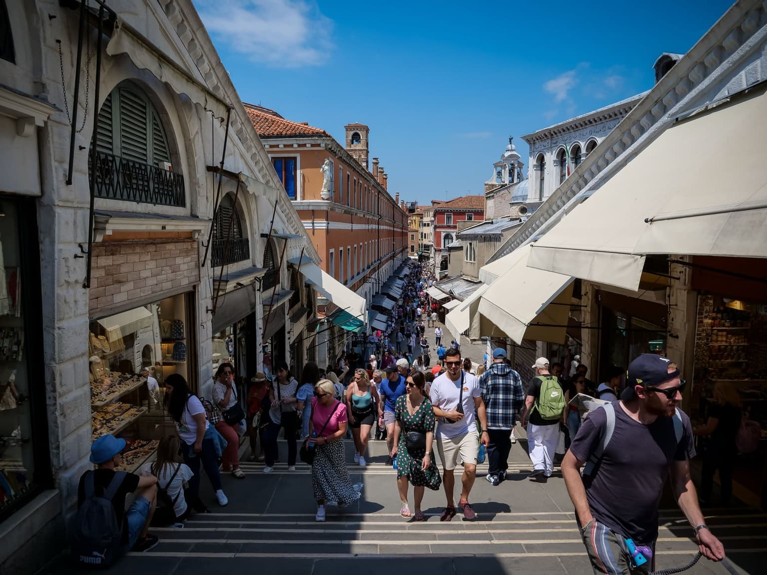 Crowds of visitors climb and descend the wide stone steps of the Rialto Bridge in Venice between rows of shops with white awnings, under a bright blue sky