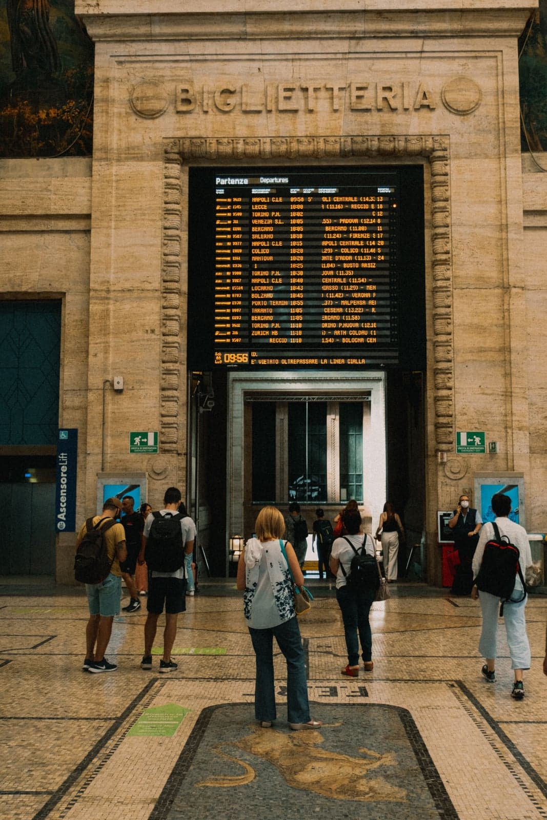 Travelers with backpacks stand on a mosaic floor in an Italian railway station hall, reading a large amber departures board set into a carved stone arch marked Biglietteria, with Venezia among the listed destinations