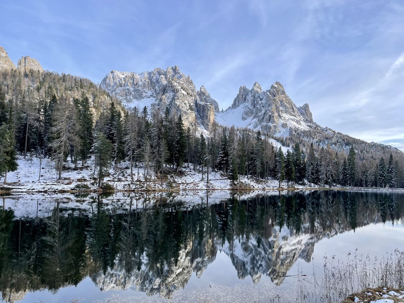 A snow-dusted Dolomite peak mirrored in a still alpine lake in winter, pine forest along the shore