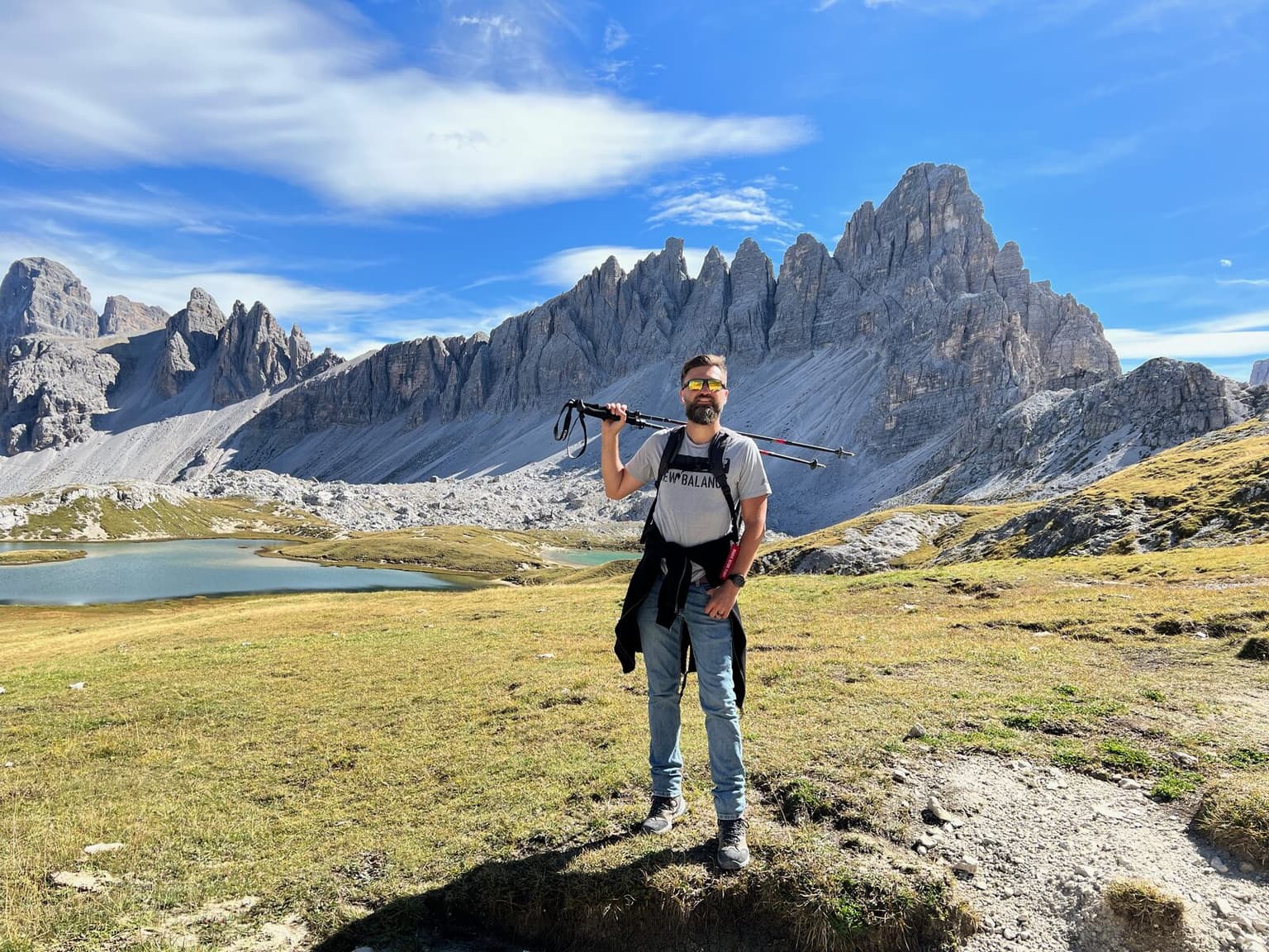 George Mandrovny on a hiking trail below the Tre Cime di Lavaredo in the Dolomites, trekking poles over his shoulder, the Laghi dei Piani lakes behind him