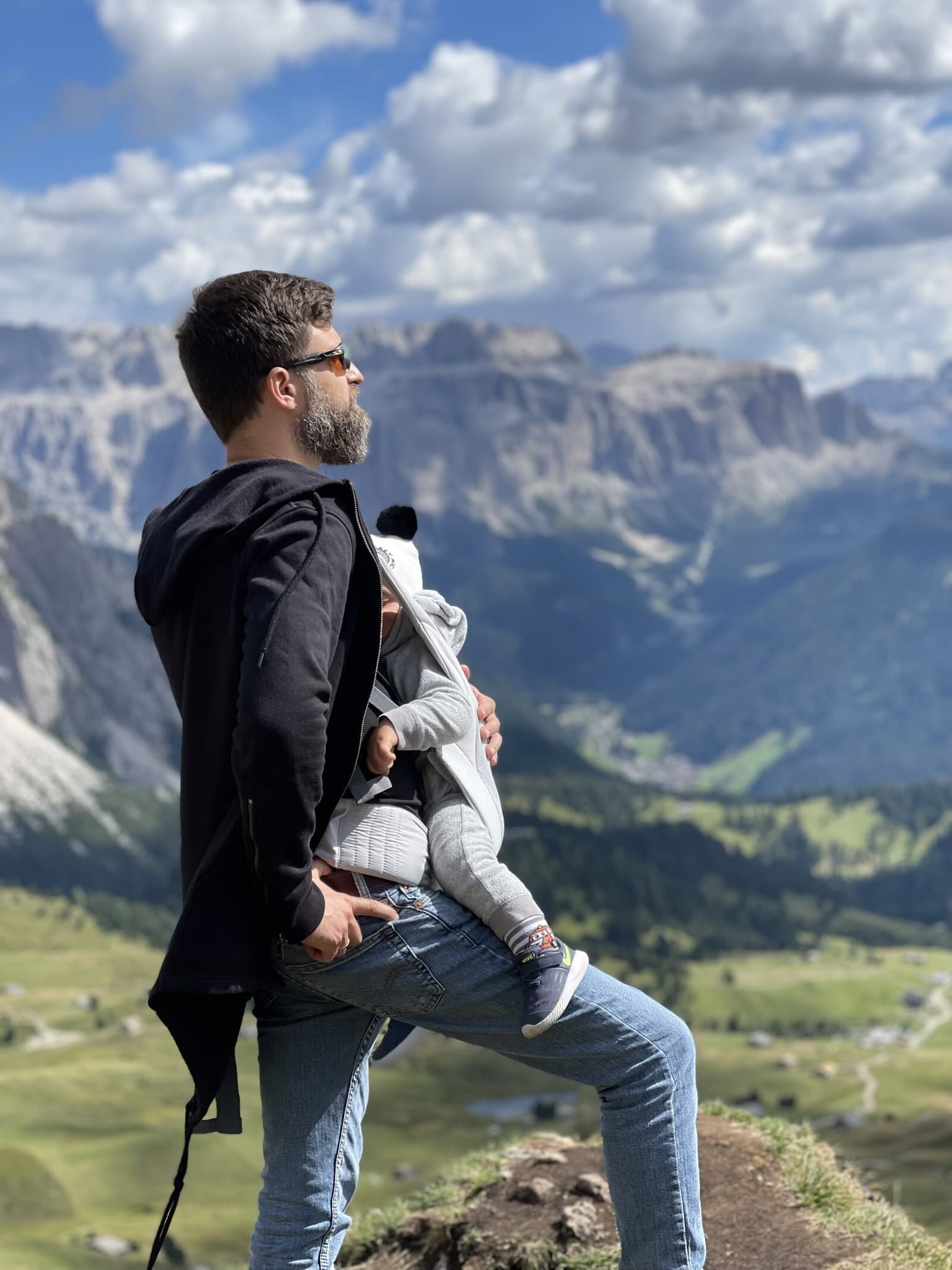 George Mandrovny carrying his infant son in a chest carrier on a ridge above a green valley, the Sella massif of the Dolomites behind him