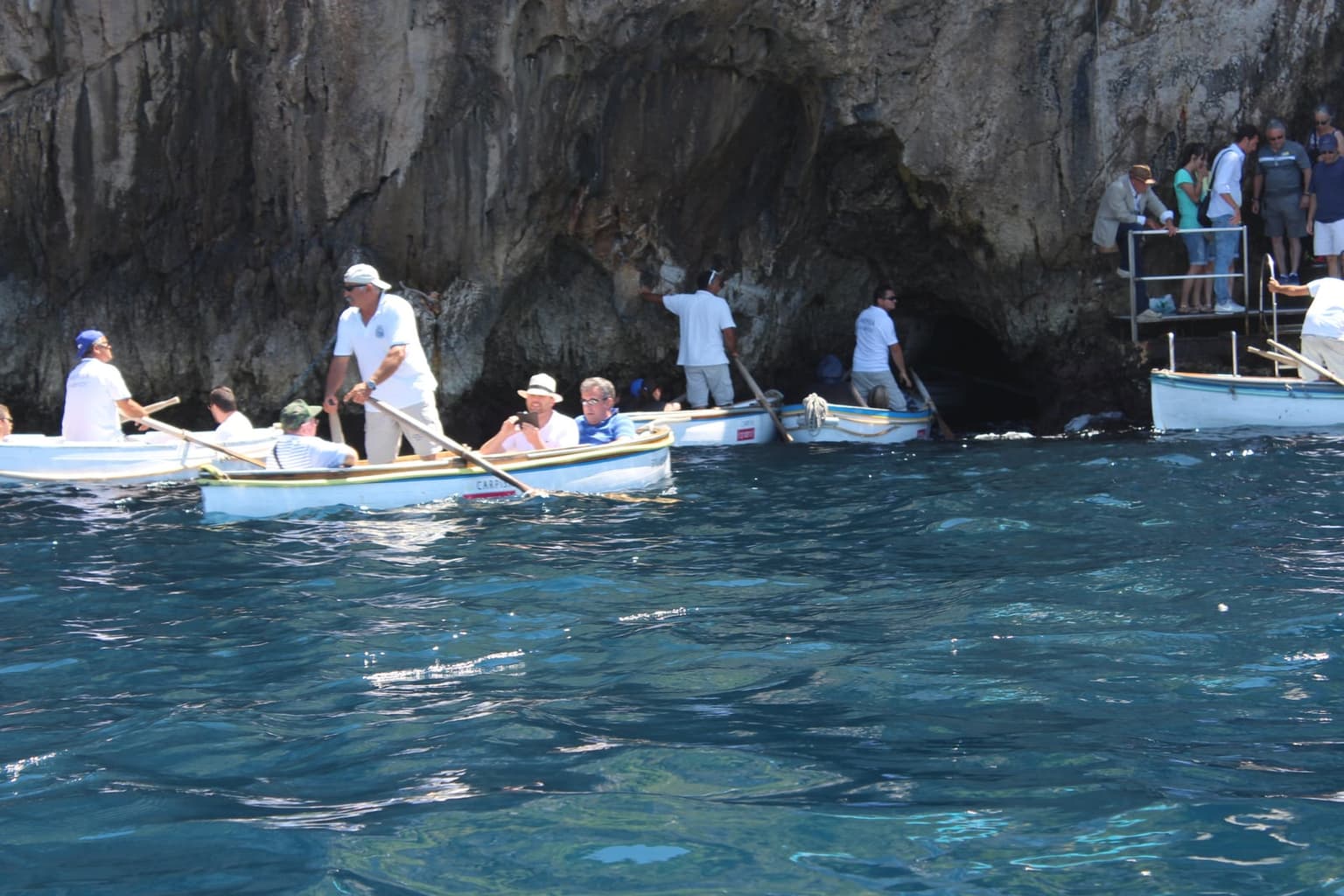 Boatmen in white shirts steering small rowboats of visitors to the low, dark entrance of the Blue Grotto at the foot of a sheer cliff, while more visitors wait on metal steps on the rock at right