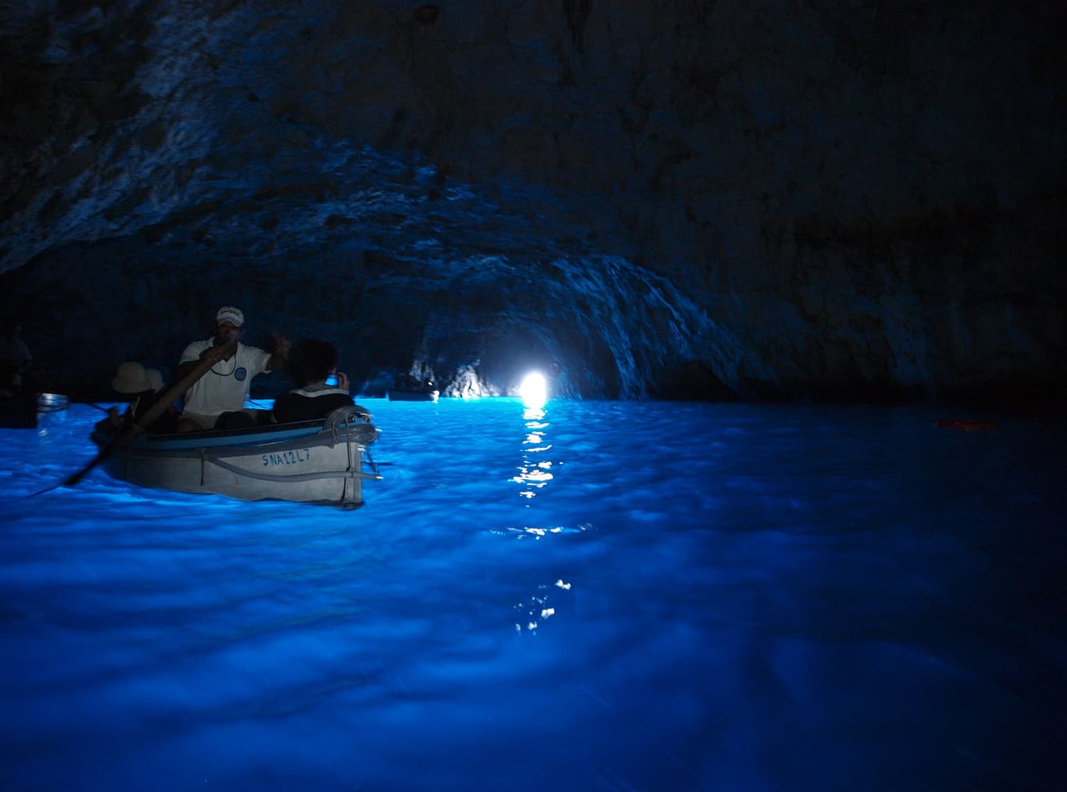 Inside the Blue Grotto, the water glows electric blue under a dark rock vault as a boatman rows a small boat of visitors, the bright entrance low on the far wall
