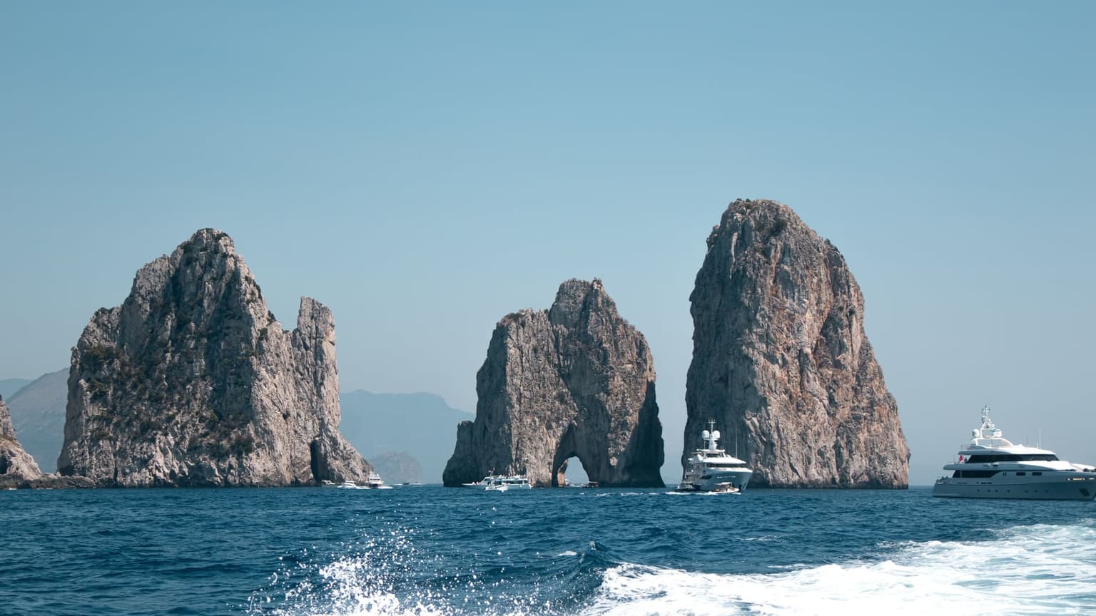 The three Faraglioni rock stacks off Capri seen from a moving boat, its wake churning in the foreground, with small boats passing through the arch in the middle stack and motor yachts idling nearby