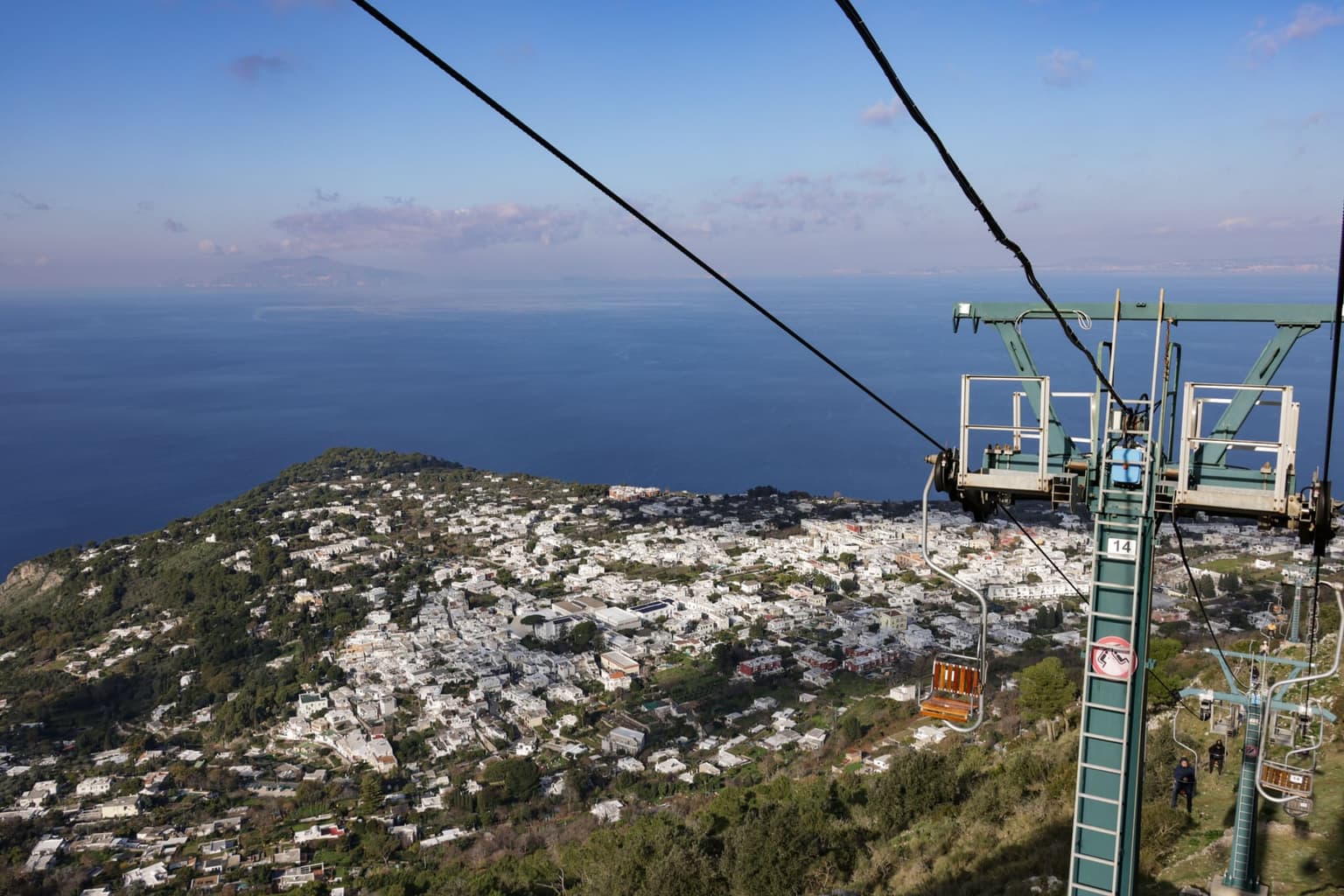 The Monte Solaro chairlift seen from a chair, an empty single wooden seat hanging beside a green pylon above the slope, with the white houses of Anacapri spread out below and the open sea beyond