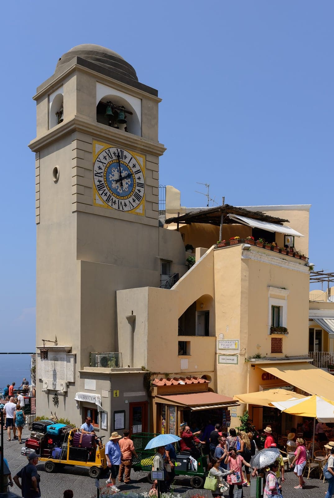 The clock tower above the Piazzetta in Capri town, with yellow cafe umbrellas and wicker chairs at its foot, a porter's cart stacked with suitcases and a summer crowd crossing the square