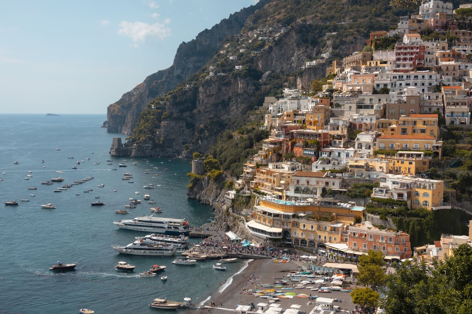 Two passenger ferries at the jetty off Positano's Spiaggia Grande, a crowd waiting on the pier and rowboats lining the dark-sand beach, while the town's ochre and pink houses climb the cliff behind