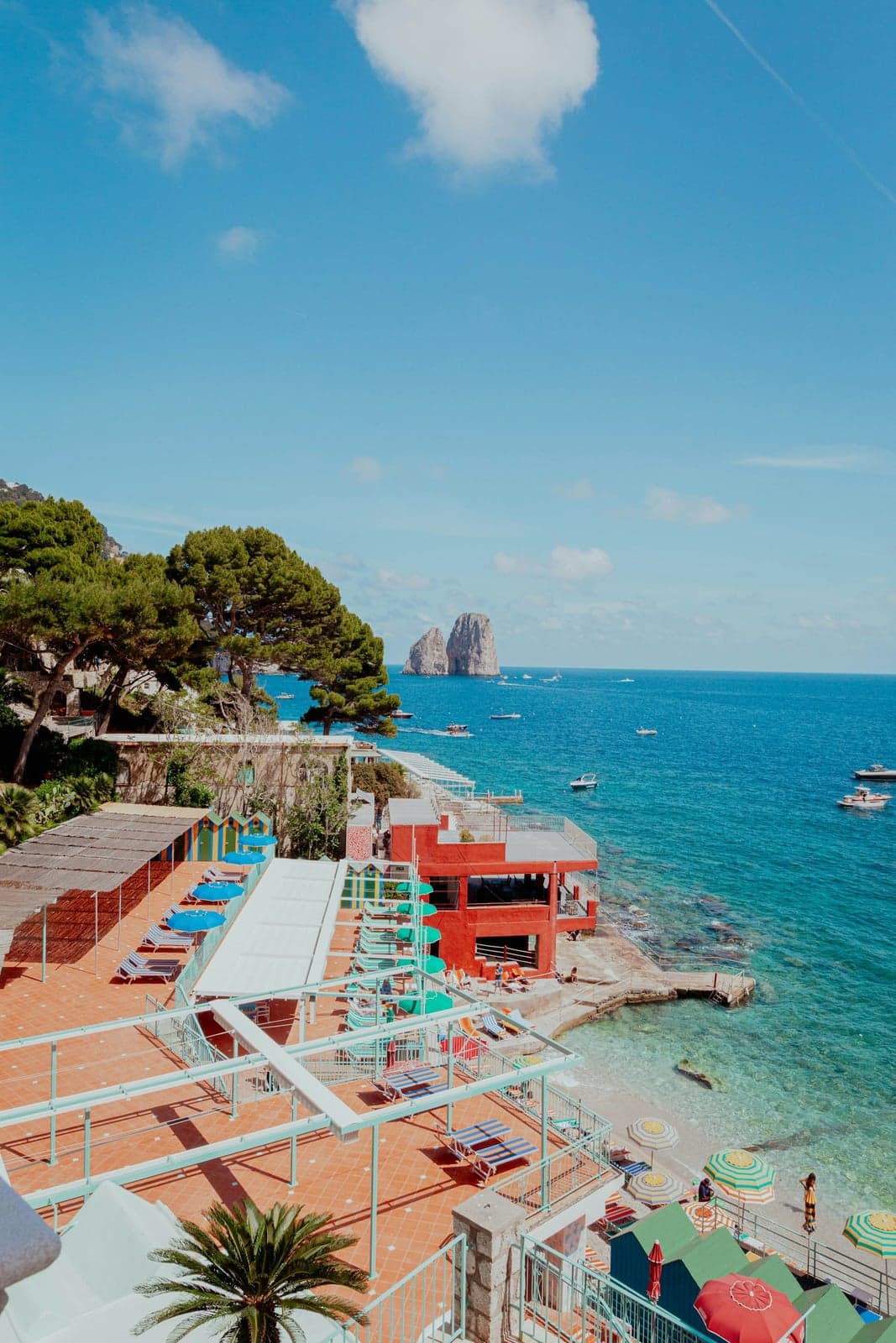 Beach-club sun decks with loungers and umbrellas and a red clubhouse above a narrow pebble beach and turquoise water on Capri's south shore, the Faraglioni stacks out at sea