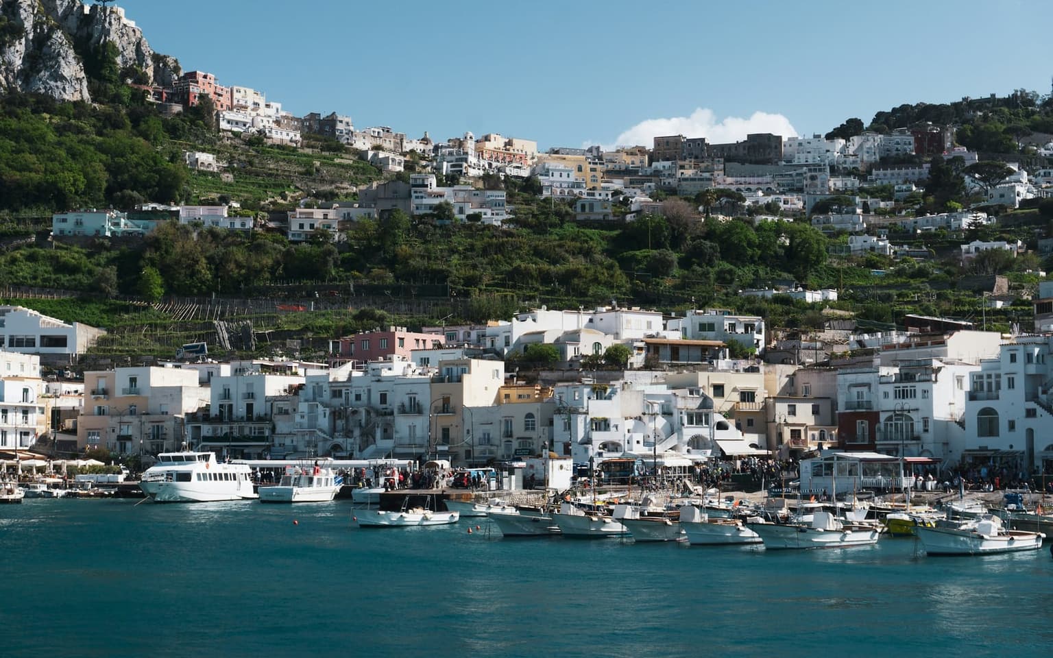 Capri's Marina Grande harbor seen from the water, white ferries and small motorboats moored along the quay below white and pastel houses that climb the green terraced hillside toward Capri town