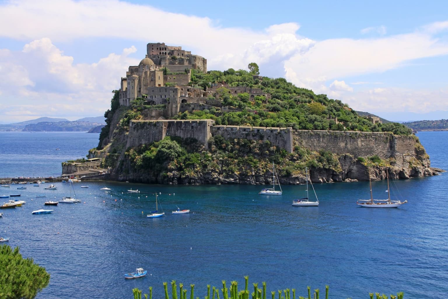 The Castello Aragonese on its rocky islet off Ischia Ponte, a domed church and fortress walls above green terraces, with the low causeway at its foot and sailboats anchored in front