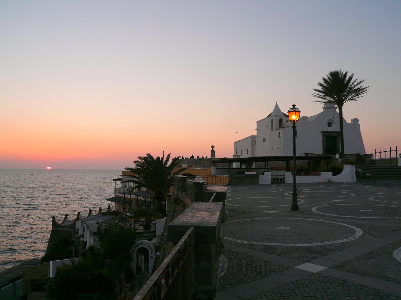Sunset from the Chiesa del Soccorso in Forio, the whitewashed church and a palm above a paved square, with the sun on the sea horizon beyond the parapet