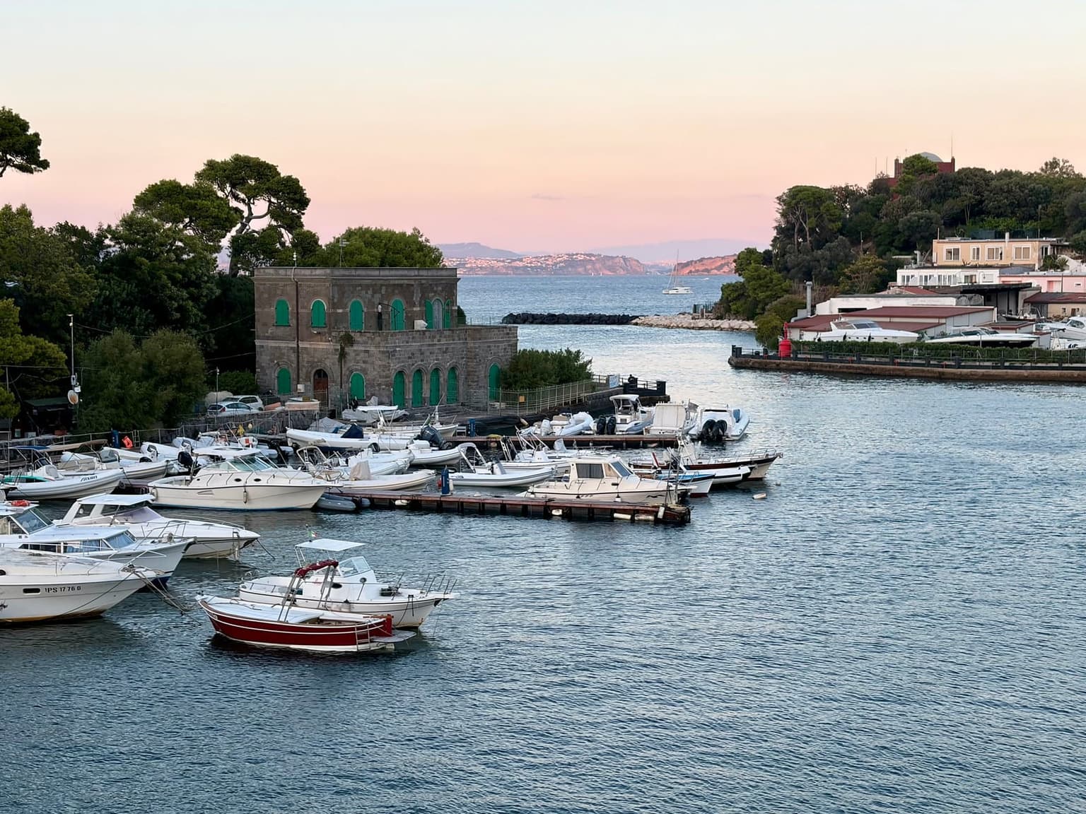 Ischia Porto's harbor at dusk, motorboats moored at floating pontoons by an old stone building, with the narrow channel leading out past a red light to the open sea