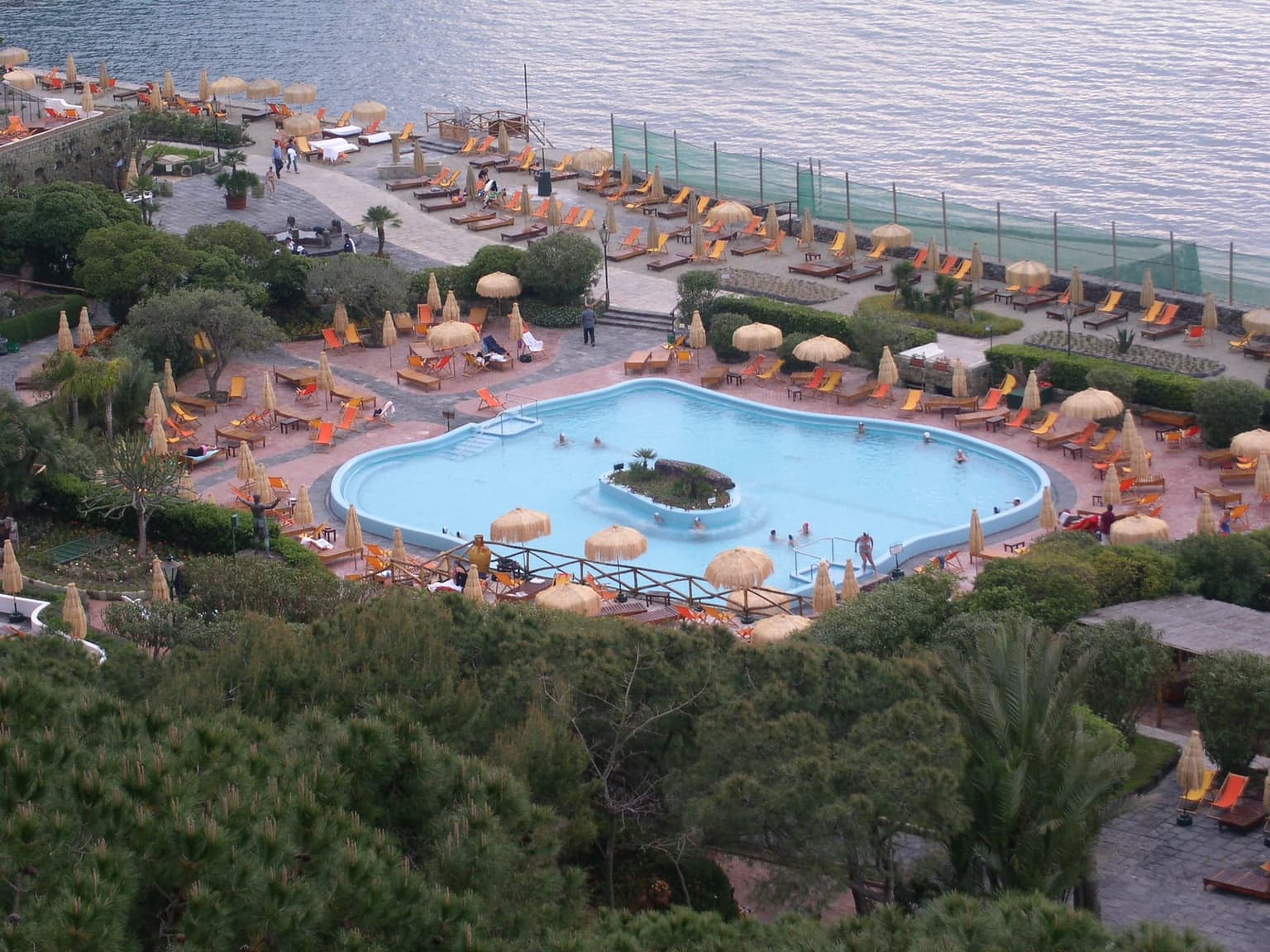 A thermal park on Ischia seen from above, a curved pool around a small planted island, straw umbrellas and loungers on the terraces, and the sea beyond