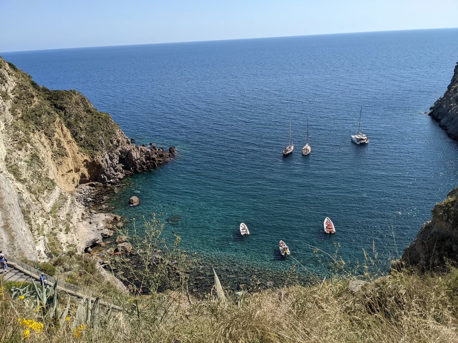 The Sorgeto cove from the cliff top, clear turquoise water over a stony shore, boats at anchor, and the long staircase down the cliff at lower left
