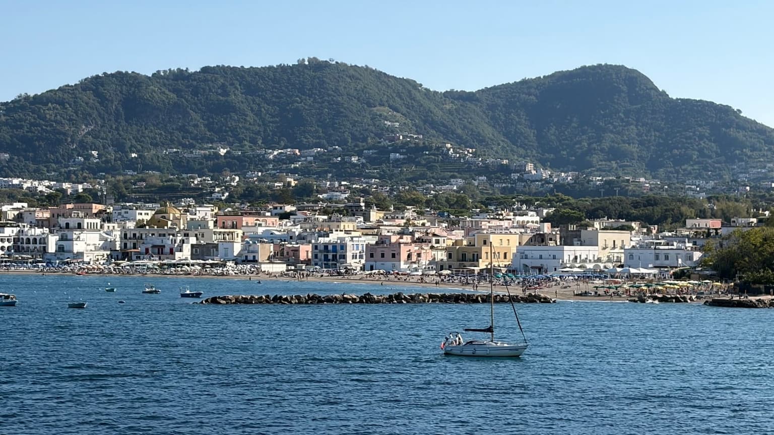 Ischia seen from the sea, a beachfront of white, pink and yellow buildings with a church dome under steep green hills dotted with houses, and a sailboat anchored in front