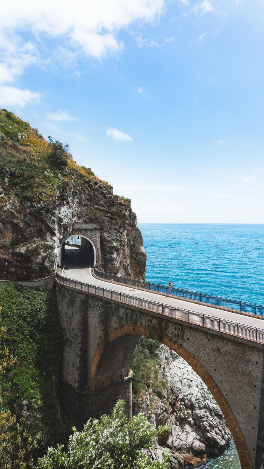 The Amalfi Coast road crossing a stone arch bridge over the Furore fjord and running into a tunnel cut through the cliff, open blue sea beside it