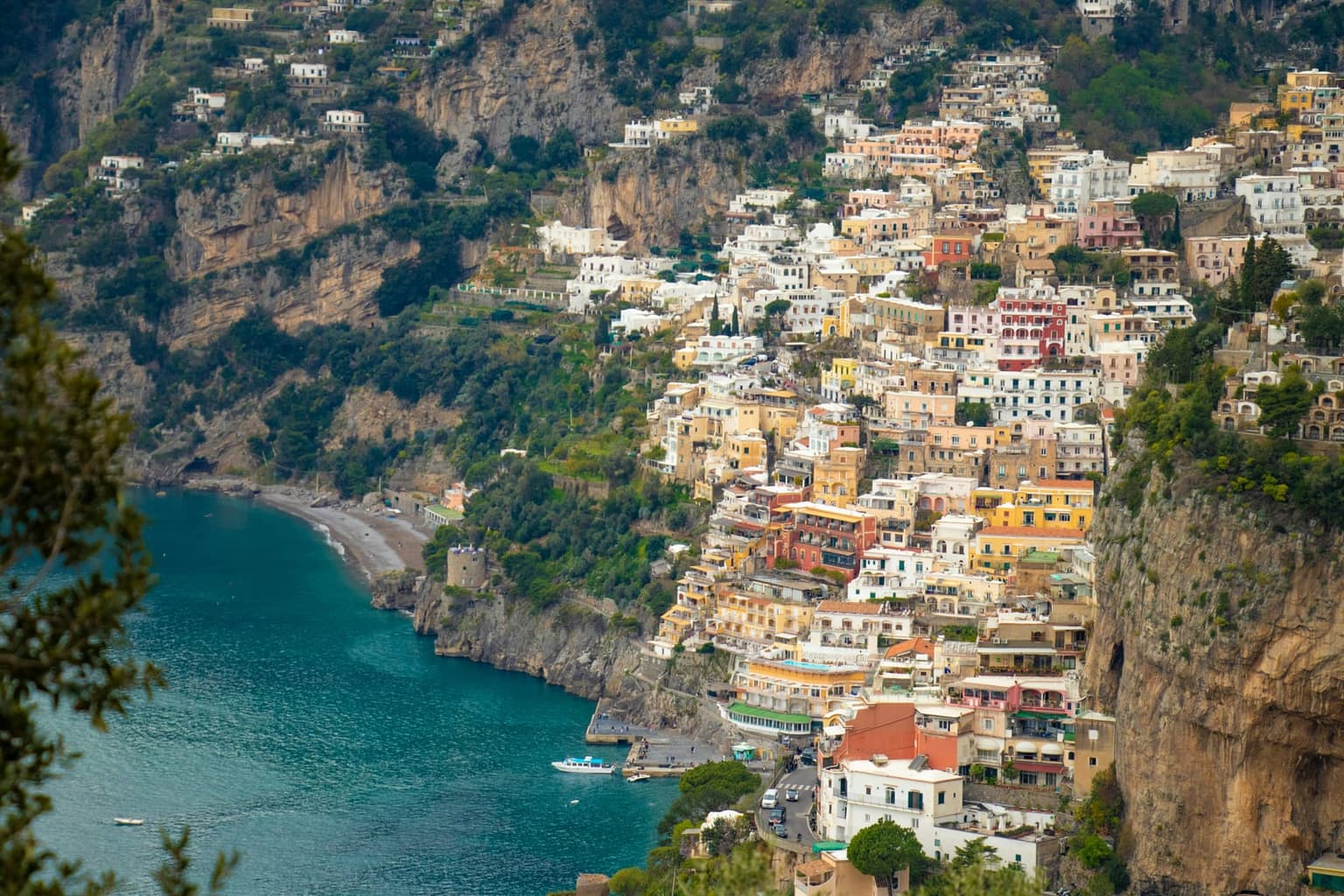 Positano seen from high on the hillside to the west, houses stacked up the cliff above the small pier, the curve of Fornillo beach and a round stone watchtower on the point