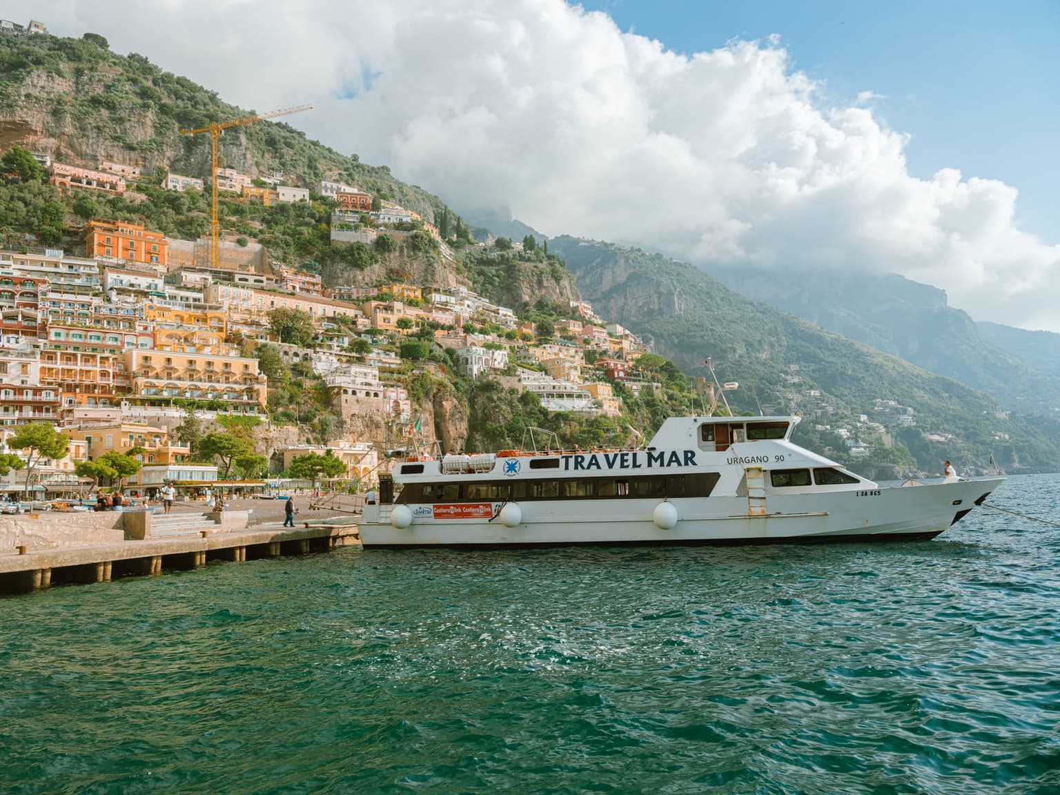 A white Travelmar ferry tied up at the concrete landing stage in Positano, the town's pastel houses climbing the cliff behind the pier and the coast fading into cloud