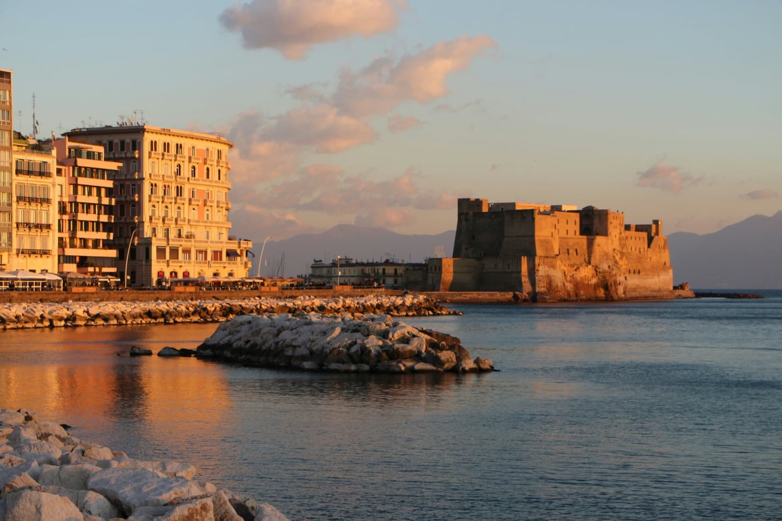 Castel dell'Ovo at sunset from the Santa Lucia seafront, with hotels along the waterfront and rock breakwaters in the bay