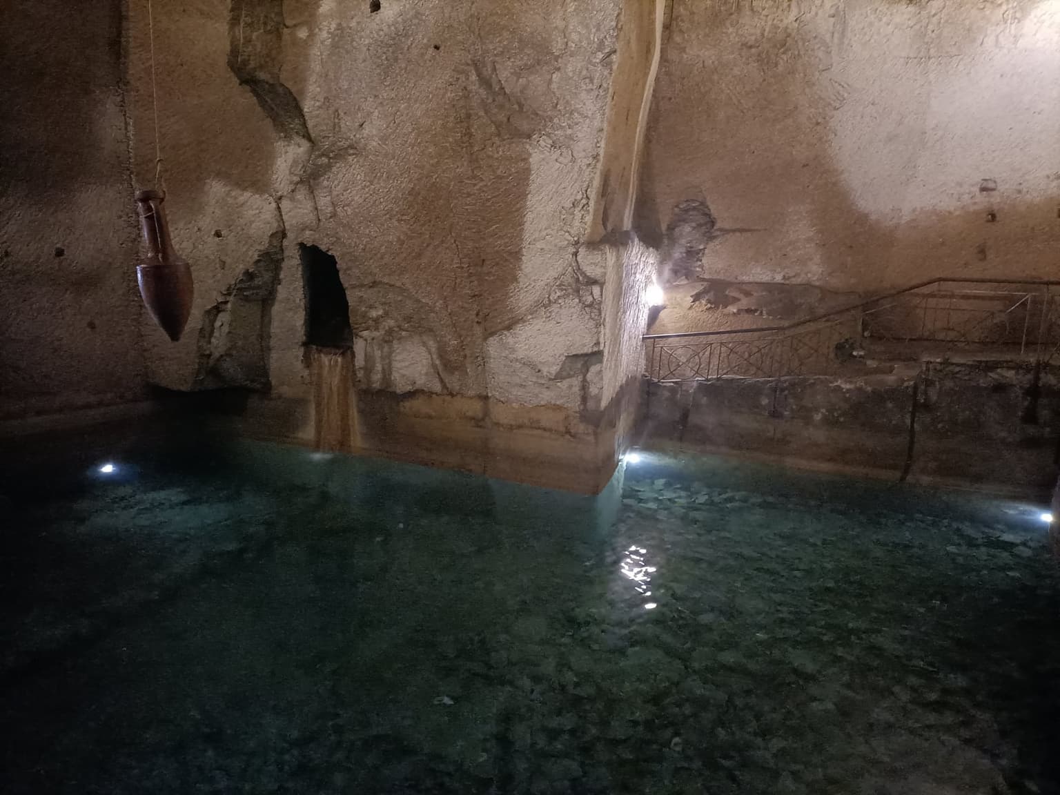 A lit cistern carved into yellow tuff in Napoli Sotterranea, with a clay amphora hanging over the clear water