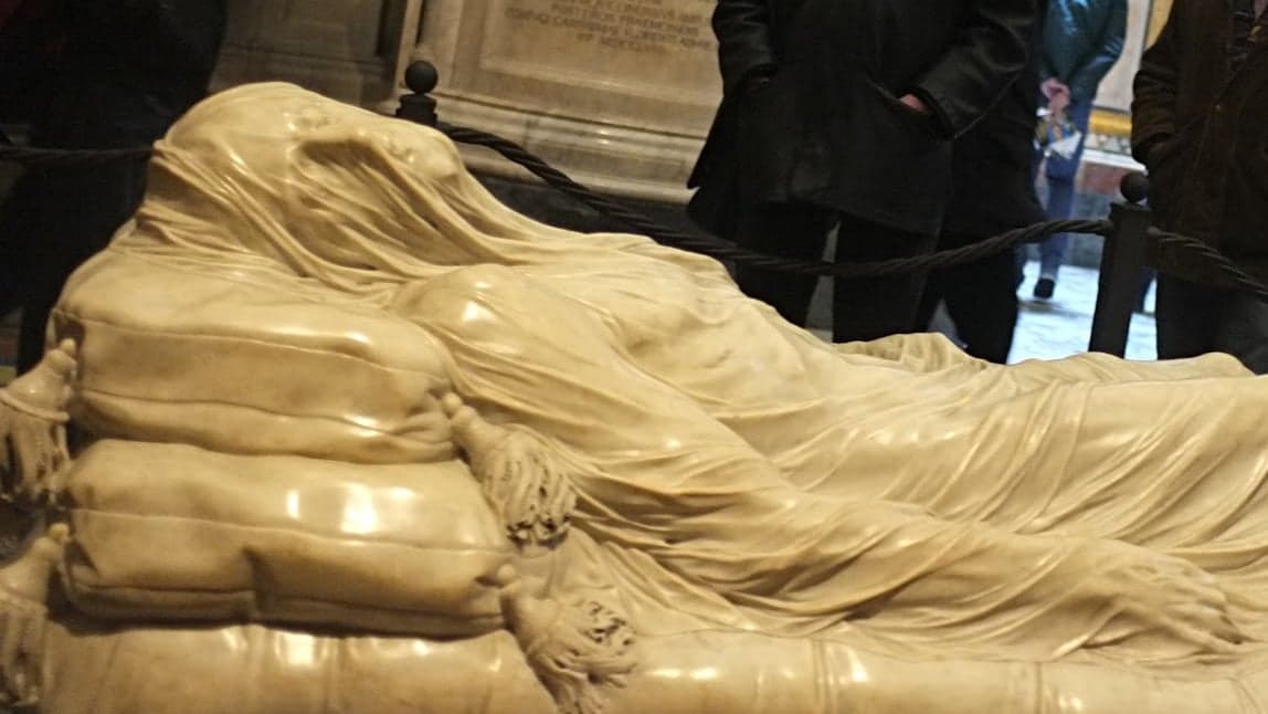 Giuseppe Sanmartino's Veiled Christ in the Cappella Sansevero, the marble figure lying on two tasseled cushions under a thin carved veil that clings to the face, chest, arms and legs, with visitors standing behind a rope barrier