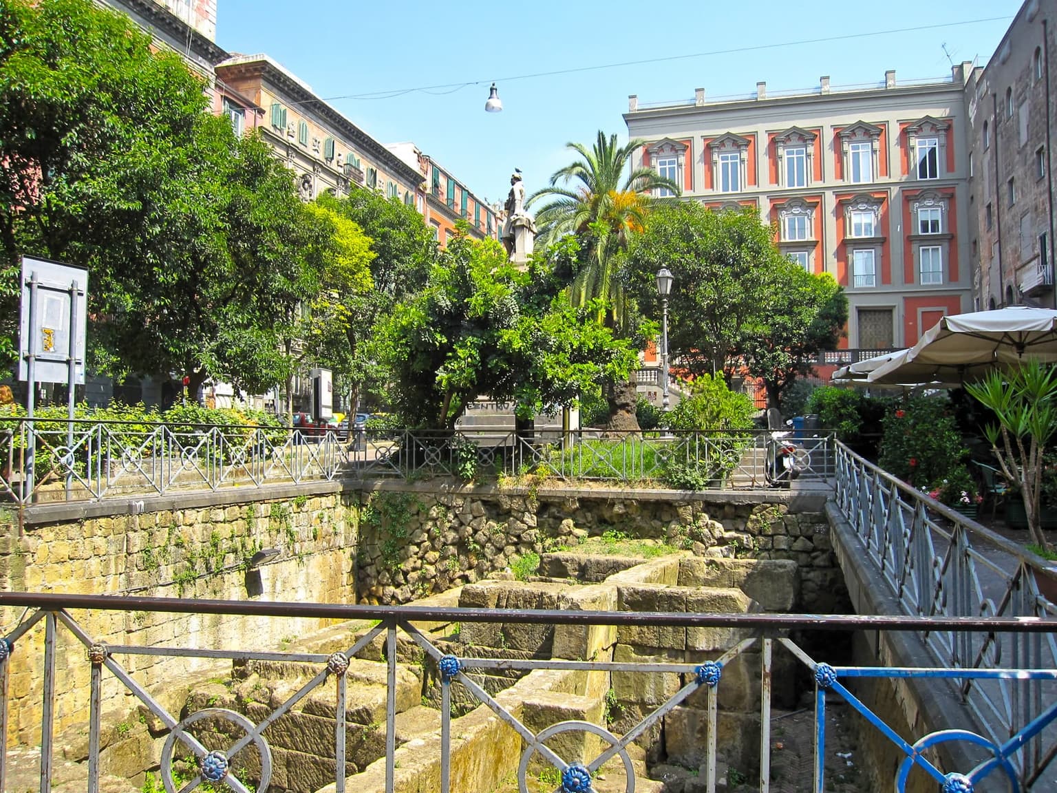 Piazza Bellini, with the excavated stones of the ancient Greek city walls below a railing, trees around a statue and cafe umbrellas