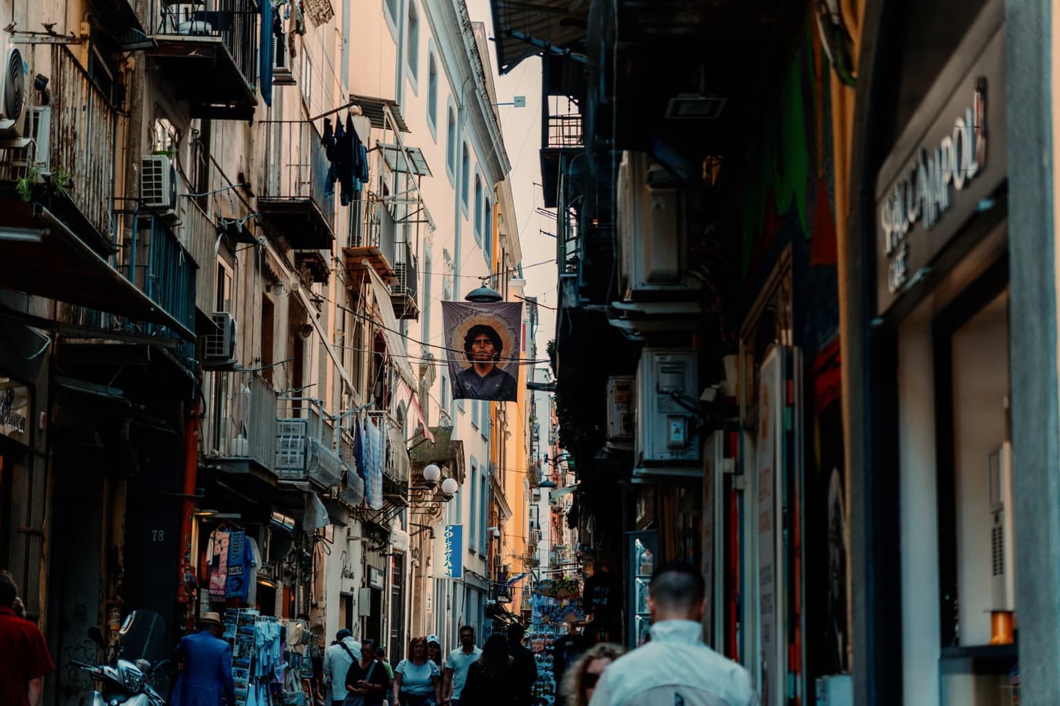 A narrow lane in Naples' historic center, with laundry on the balconies, a Maradona banner strung overhead, a parked scooter and souvenir stalls as people walk through