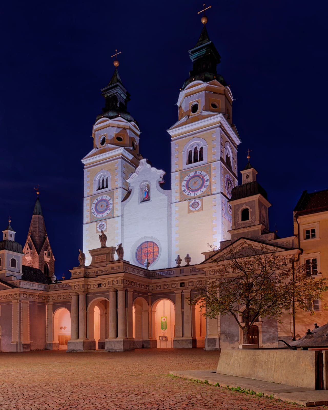 The twin clock towers and white facade of Bressanone cathedral lit up at night above the empty cobbled Piazza Duomo, with an arcaded portico along its base