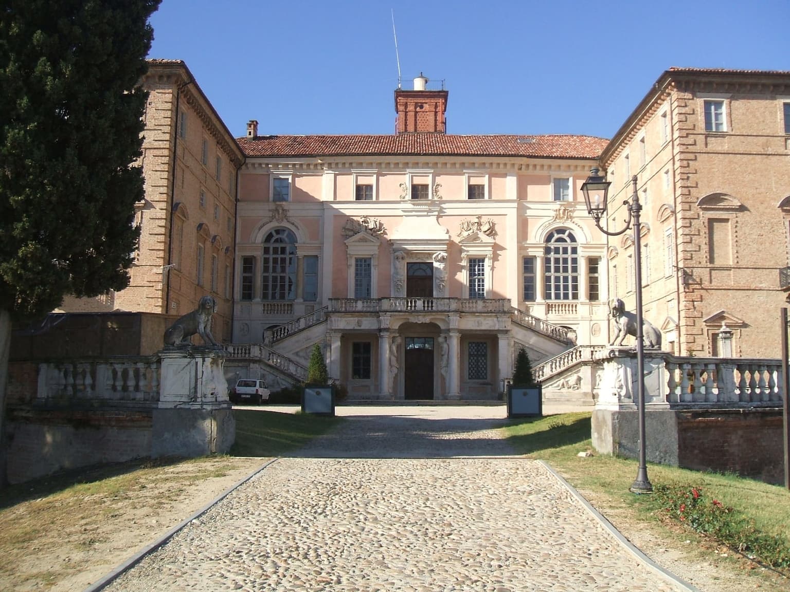 The pink facade of Govone's castle with a double stone staircase, seen up a cobbled drive between stone lions on the gateposts