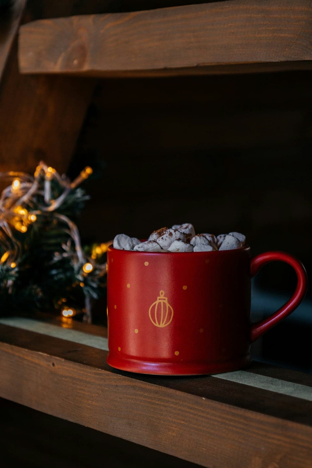 A red Christmas mug with a gold ornament print, filled with a hot drink topped with marshmallows and a dusting of cinnamon, on a wooden shelf beside a sprig of fir and fairy lights