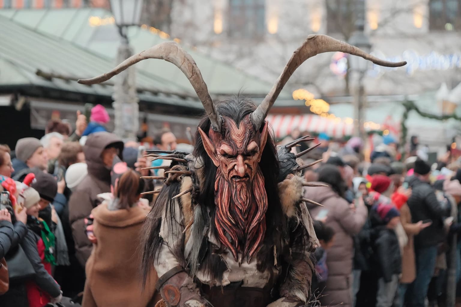 A Krampus in a carved red wooden mask with long curved horns, a shaggy fur costume and spiked shoulders, walking through a crowd of onlookers holding up phones, Christmas market stalls blurred behind