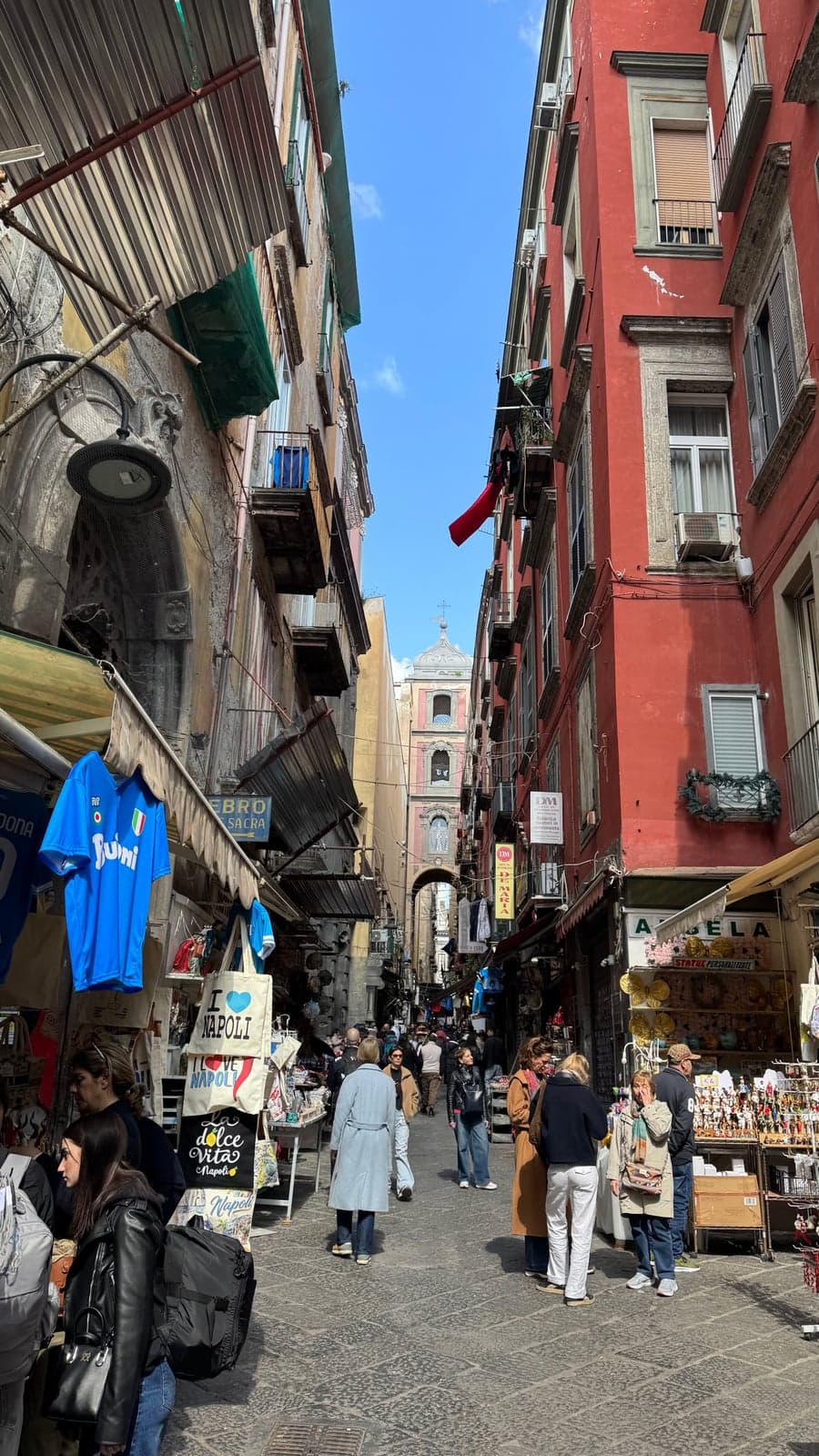 Narrow Via San Gregorio Armeno in Naples, lined with shop fronts selling nativity figurines and souvenirs under tall red and ochre buildings, with an arched bell tower over the street at the far end