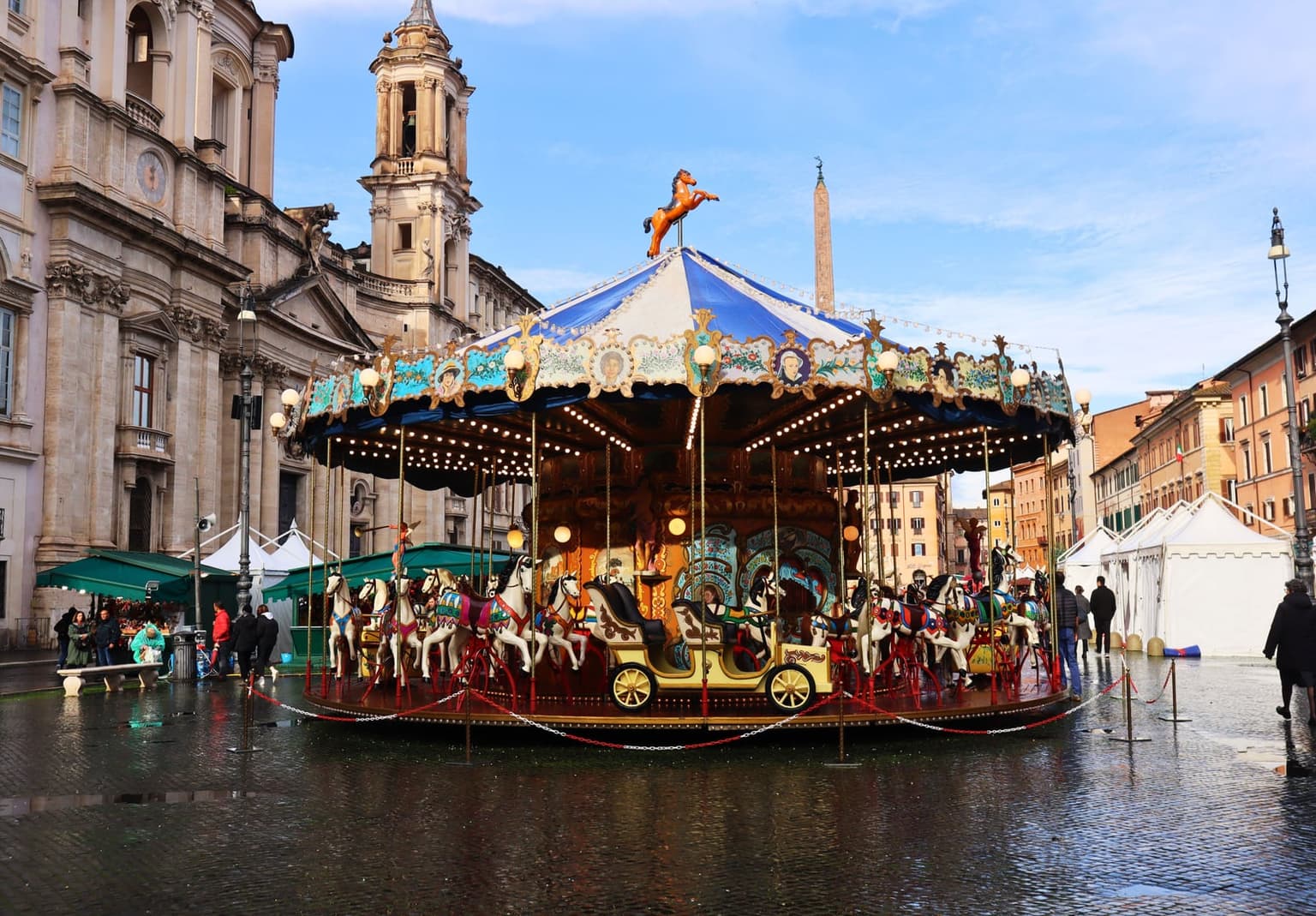 A vintage carousel with painted horses on rain-wet Piazza Navona, the bell tower of Sant'Agnese in Agone and the obelisk behind it, fair stalls with green awnings and white tents along the edges