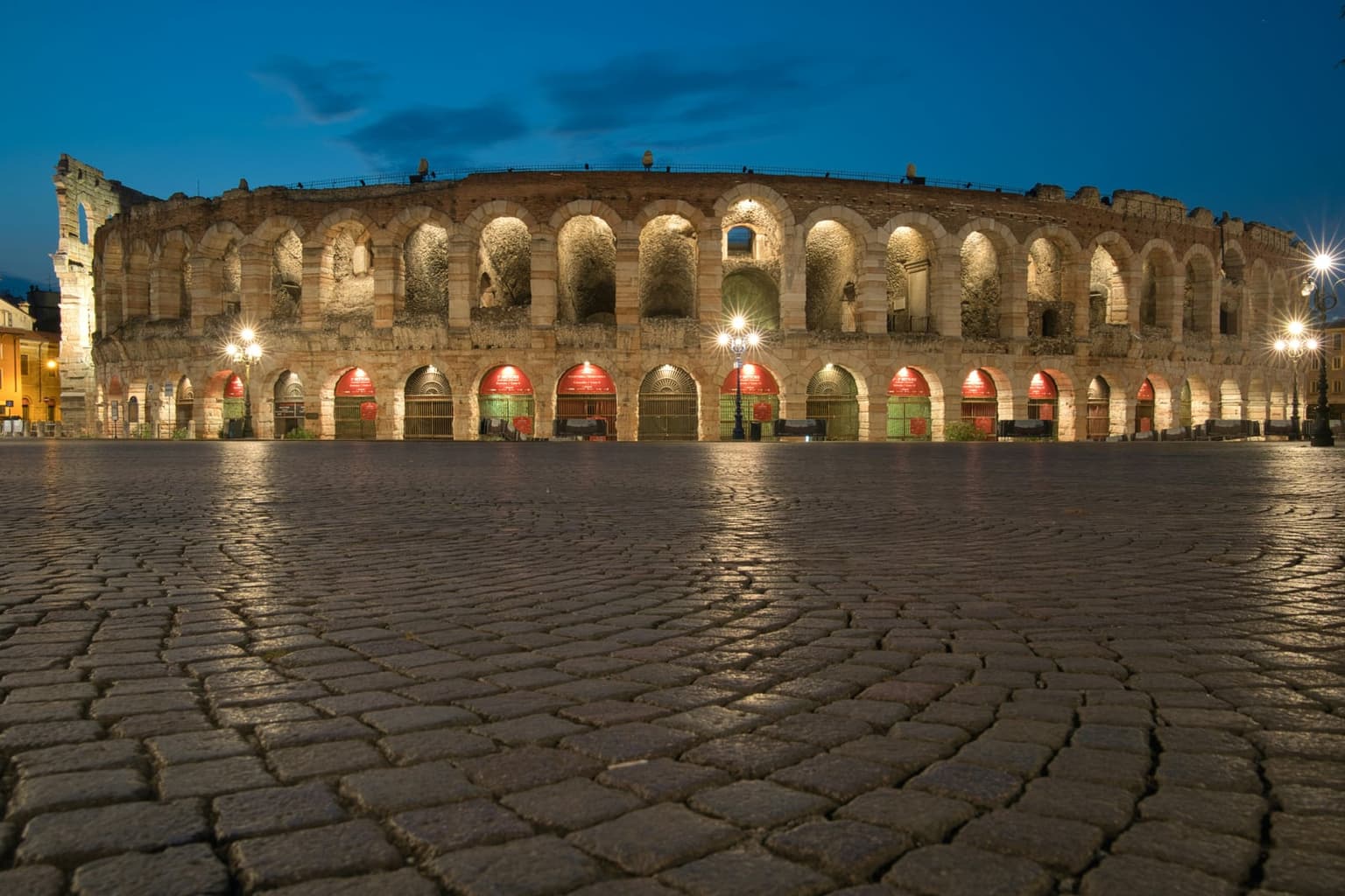 Verona's Roman Arena at blue hour, its two tiers of stone arches lit warm and the ground-floor arches glowing red, behind an empty cobbled Piazza Bra